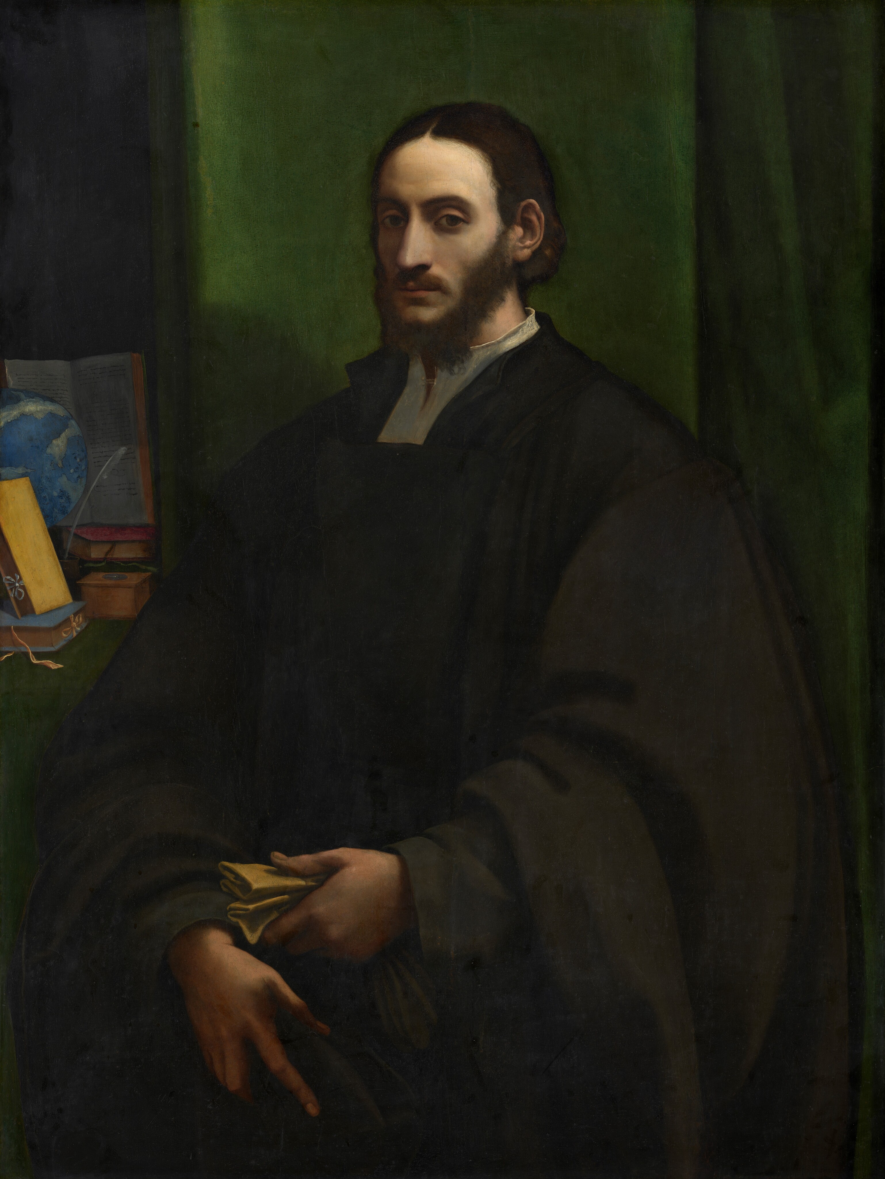 A bearded, pale-skinned man with shoulder-length, dark brown hair is shown from the hips up against a dark, emerald-green curtain in this vertical portrait painting. The man’s body is angled slightly to our left, while his head turns to look directly at us with dark, shadowed eyes. His chin-length hair is parted down the middle and tucked behind his ears. He wears a dark robe over a white shirt with a high collar. In his left hand, to our right, he holds a folded piece of yellowish paper or cloth, and his other hand rests just below it with fingers pointing downward. He is lit from our right so the far side of his face is thrown into shadow. At his shoulder and to our left, a shelf or table covered with green holds a globe, an open book with pages filled with text, a stack of closed books, and a quill resting in an inkwell. Some of the text in the open book is visible but not clearly legible. Most of the background is filled with the jewel-toned green curtain, but a sliver to the left, behind the objects, is black.