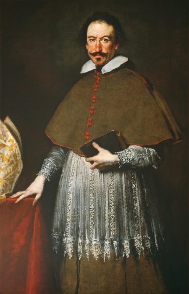 This painting shows a man from the legs up, his body facing slightly to the left but his head turning to look at us. He has pale skin, ruddy cheeks, short, dark hair, a dark, curling mustache, a dark goatee, arched eyebrows, deep-set eyes, and thin lips set in a neutral expression. He wears a dark green smock with a white lace collar, and beneath that a white lacy garment over a dark green robe. He holds a black book in his left hand, and his right hand rests on a table next to him that is draped in red fabric. On the table appears to be a tall white hat with gold detailing. The background is a dark brown-green color.