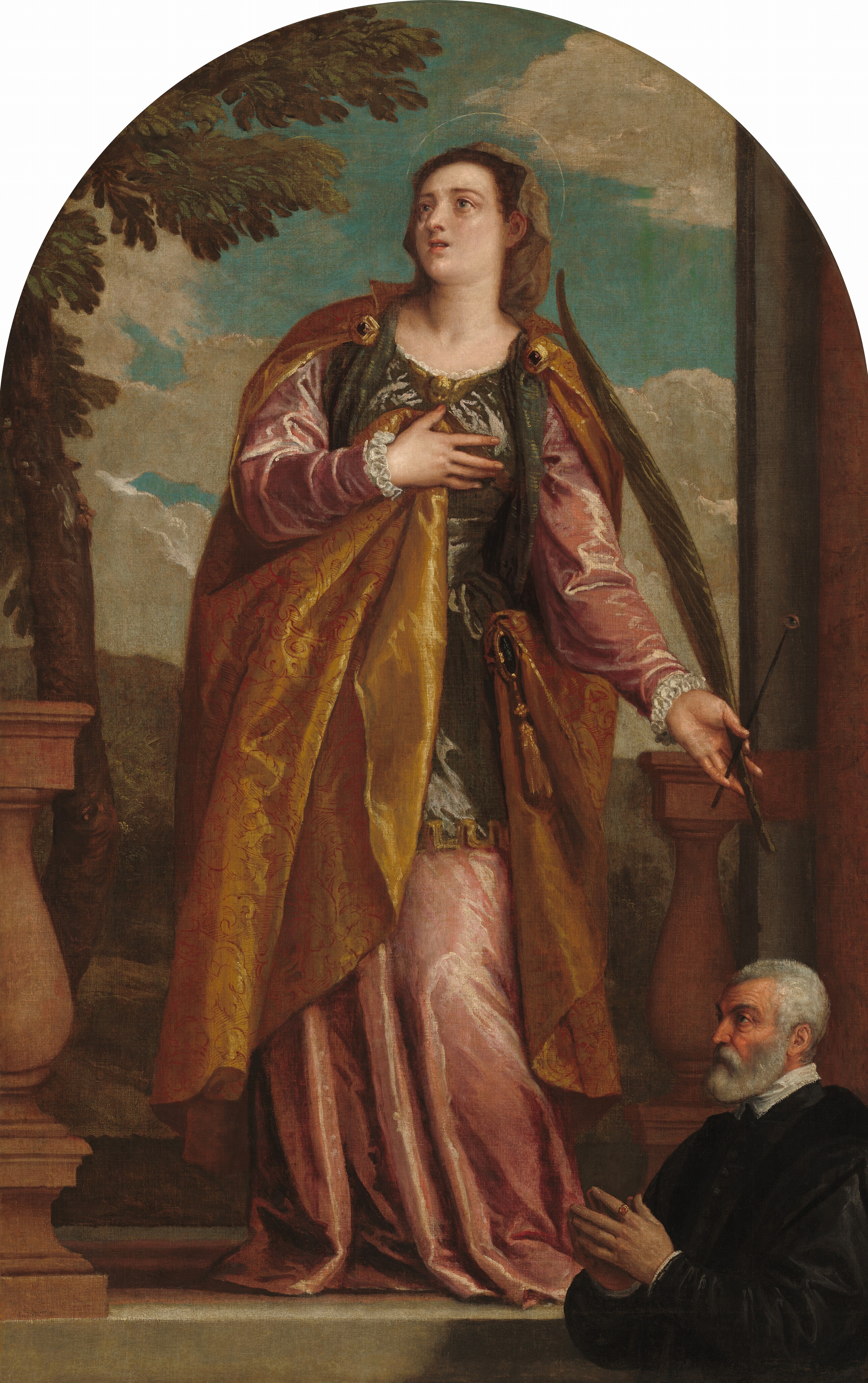 A woman with a delicate, translucent halo stands on a stone ledge above an older man who is shown in a smaller scale in the lower right of this vertical, arched painting. Both have pale skin. The woman faces us with her right hand, on our left, pressed to her chest. Her head is turned to look up and to the left with wide, gray eyes under thin brows. Her left knee, on our right, is slightly bent, making her torso lean in that direction. She has a round face with a long nose and a small coral-red mouth, which is open. Her brown hair is covered by a sable-brown veil, and she wears a sleeveless olive-green tunic over a long-sleeved dusty rose-pink gown. The tunic is adorned with a gold, winged baby head at the neckline. A copper-red cloak covered with a delicate cherry-red pattern is fastened to the shoulders of her tunic with gold ornaments embedded with dark red gems. The cloak wraps around her right side, on our left, and her right hand holds a fold of the cloak to her chest. The left side of the cloak wraps around that hip and is attached with a round, gold object with a tassel hanging from the bottom. She carries a dark green palm frond in her left hand, along with a short staff topped with a human eye. She stands in a gap between terracotta-colored balustrades with brown hills and a cloud-filled, teal-blue sky beyond her. The man in the lower right stands facing our left, almost in profile as if in front of the ledge on which the woman stands. He has a ruddy, deeply lined face, short gray hair and a trimmed beard, and wears a voluminous black coat with a white collar. The fingertips of his raised hands touch and a gold ring with a burnt-orange gem is on the first finger of his near hand.