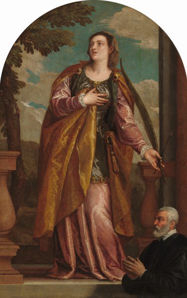 A woman with a delicate, translucent halo stands on a stone ledge above an older man who is shown in a smaller scale in the lower right of this vertical, arched painting. Both have pale skin. The woman faces us with her right hand, on our left, pressed to her chest. Her head is turned to look up and to the left with wide, gray eyes under thin brows. Her left knee, on our right, is slightly bent, making her torso lean in that direction. She has a round face with a long nose and a small coral-red mouth, which is open. Her brown hair is covered by a sable-brown veil, and she wears a sleeveless olive-green tunic over a long-sleeved dusty rose-pink gown. The tunic is adorned with a gold, winged baby head at the neckline. A copper-red cloak covered with a delicate cherry-red pattern is fastened to the shoulders of her tunic with gold ornaments embedded with dark red gems. The cloak wraps around her right side, on our left, and her right hand holds a fold of the cloak to her chest. The left side of the cloak wraps around that hip and is attached with a round, gold object with a tassel hanging from the bottom. She carries a dark green palm frond in her left hand, along with a short staff topped with a human eye. She stands in a gap between terracotta-colored balustrades with brown hills and a cloud-filled, teal-blue sky beyond her. The man in the lower right stands facing our left, almost in profile as if in front of the ledge on which the woman stands. He has a ruddy, deeply lined face, short gray hair and a trimmed beard, and wears a voluminous black coat with a white collar. The fingertips of his raised hands touch and a gold ring with a burnt-orange gem is on the first finger of his near hand.