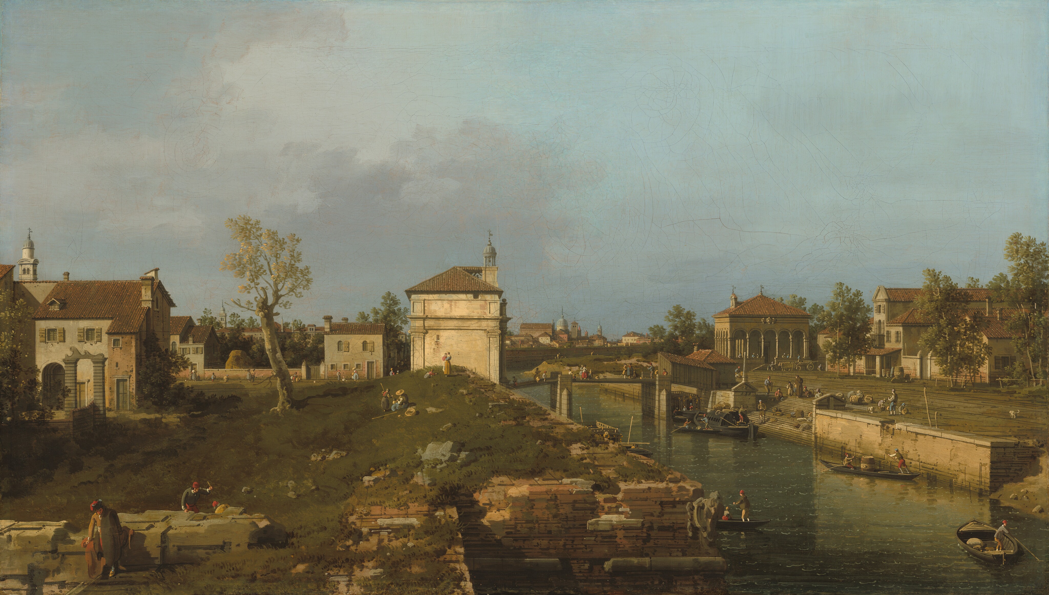 Warm light pours onto white stone and brown brick buildings in a grassy landscape in this horizontal painting. Tiny people walk, stand, sit, and work across the turf, among the buildings, and over a bridge spanning a canal to our right. The people wear long-sleeved shirts, vests, britches, stockings, dresses, hats, and caps in shades of straw yellow, olive green, apple red, muted blue, brown, and bright white. Touches of peach paint suggest they are all pale skinned. The people closest to us are three men working along what might be the remains of a stone foundation in the lower left corner. One raises an instrument while another crouches nearby on the far side of the structure, and the third leans on our side, looking down at the grass. The land rises gently back to the end of a row of structures to our left. A thick-trunked tree with a sparse canopy is halfway between those structures and a building on our side of the bridge, in the center of the composition. That bone-white structure has engaged columns with capitals, a tiled roof, and a round, gazebo-like lantern or bell tower. A brick wall between more buildings line a lane beyond the tree and these structures, and rooflines pile up along the horizon, which comes halfway up the composition. People and children stand and sit on the grassy knoll or walk along the lane. The canal angles from the lower right corner of the canvas to the center of the horizon. The walls of the canal are built with thin layers of peach and tan-colored stone. Close to us, the wall on our side jogs to our left, creating an inlet or bay. A staircase cuts into the far wall to allow access to the water. Boats steered by men using long sticks carry people or supplies. A more elaborate covered boat, likely a ferry, pulls up to the base of the stairs. The stairs lead up to a plaza, which is lined with peanut-brown buildings with terracotta-brown roofs. People and dogs mill about the square and cross the bridge beyond the ferry. Some smoky-purple clouds drift into the scene from the left against an otherwise clear blue sky.