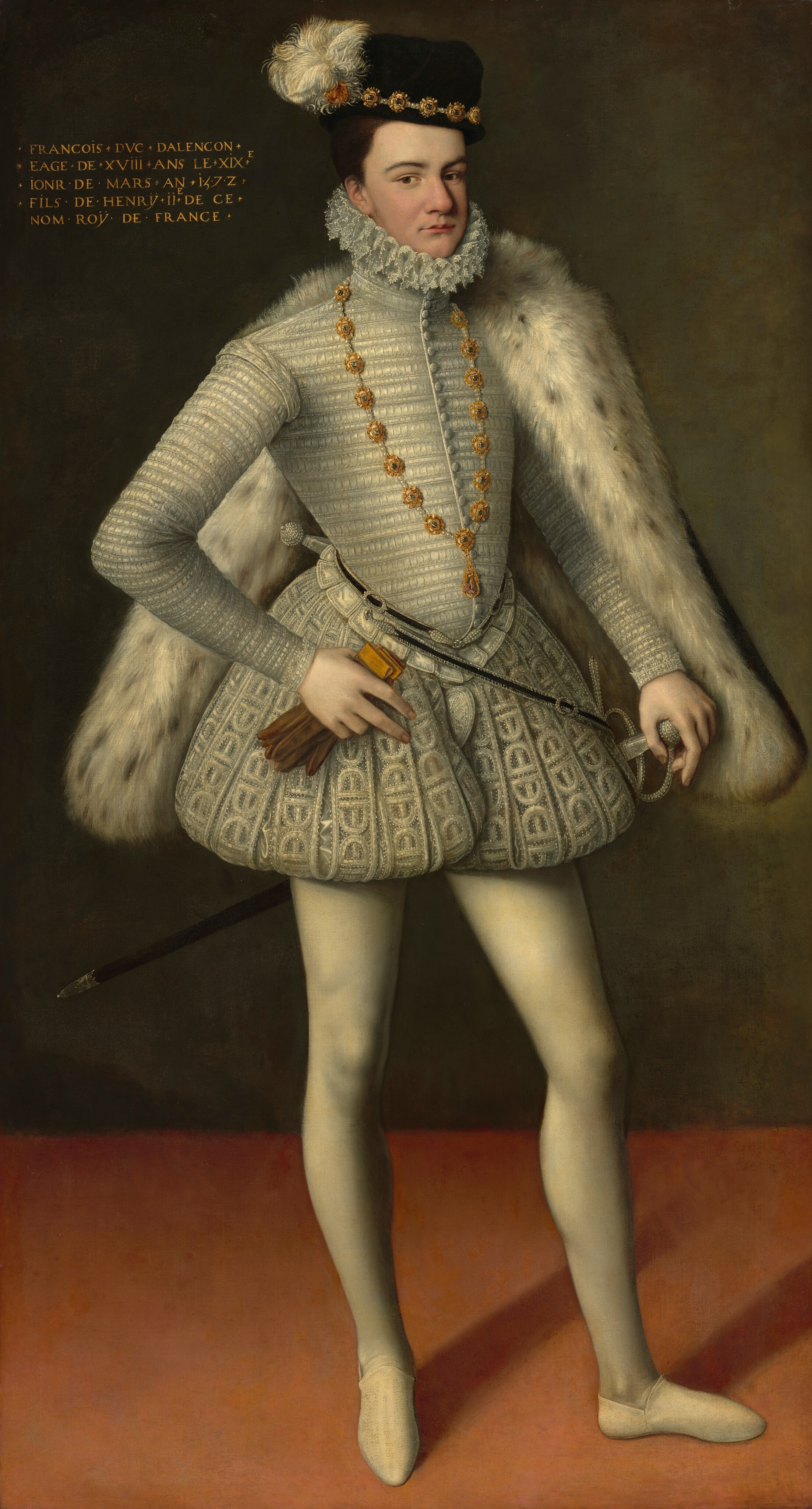 A clean-shaven, pale-skinned man wearing all white, a black-speckled white fur cape, and brimless black hat stands and looks at us in this full-length, vertical portrait painting. The man’s body and face are angled to our right, but he looks at us from the corners of his brown eyes under dark, curved brows. He has a slightly bulbous nose, the suggestion of a cleft chin, and his thin, pink lips are closed. His brown hair is cut short under the cap, which has a high crown. The hat is lined with jeweled gold medallions and pearls around the bottom edge, and the barbs of the white feather flicker like a busy flame. The lace-edged ruff, which is pleated in figure eights, hugs his chin and the back of his neck. The high-collared, white, long-sleeved jacket is textured in narrow, horizontal bands. A narrow black belt decorated with silver trim and pearls loops around his waist, and a sword hangs from the far hip. The white-on-white patterned breeches puff out at the waist and are gathered high on the thigh, like puffy shorts. He wears smooth, cream-white stockings and the same color slippers. The hip-length fur cape hooks over the far shoulder, and that hand rests on the pearled hilt of a thin sword. His other elbow is bent, and he holds a pair of gold and brown-colored gloves in that hand. The first two fingers are extended to lie along the fabric of his pants. The wall behind the man is earthy green and the floor a rosy peach. Gold text in the upper left corner of the painting reads, “FRANCOIS DVC DALENCON EAGE DE XVIII ANS LE XIXE IONR DE MARS AN 1572 FILS DE HENRŸ II DE CE NO ROŸ DE FRANCE.”
