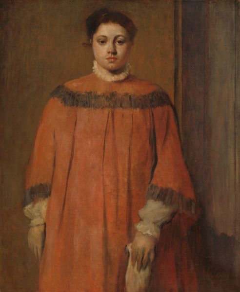 Shown from the knees up, a pale-skinned woman in a voluminous rust-orange dress stands facing us in this vertical portrait painting. She looks toward us but slightly down with dark brown eyes under curved brows. She has a wide face, rounded cheeks, a broad nose, and closed, coral-pink lips over a deeply dimpled chin. Her chestnut-brown hair is pulled up and back. The orange dress has a band of charcoal gray across the shoulders and at the flaring cuffs. Wide, vertical pleats fall from under the band across her chest. White trim, perhaps lace or fur, is suggested by loosely painted cream white at the high neck. The poofy sleeves and cinched cuffs of her white shirt extend beyond the dress sleeves. Her right arm, to our left, hangs by her side and the other holds an indistinct, loosely painted light-colored object down in front of her. She seems to be in a corner with the wall behind thinly washed in muted brown and green and a sienna-brown wood panel wall to our right.