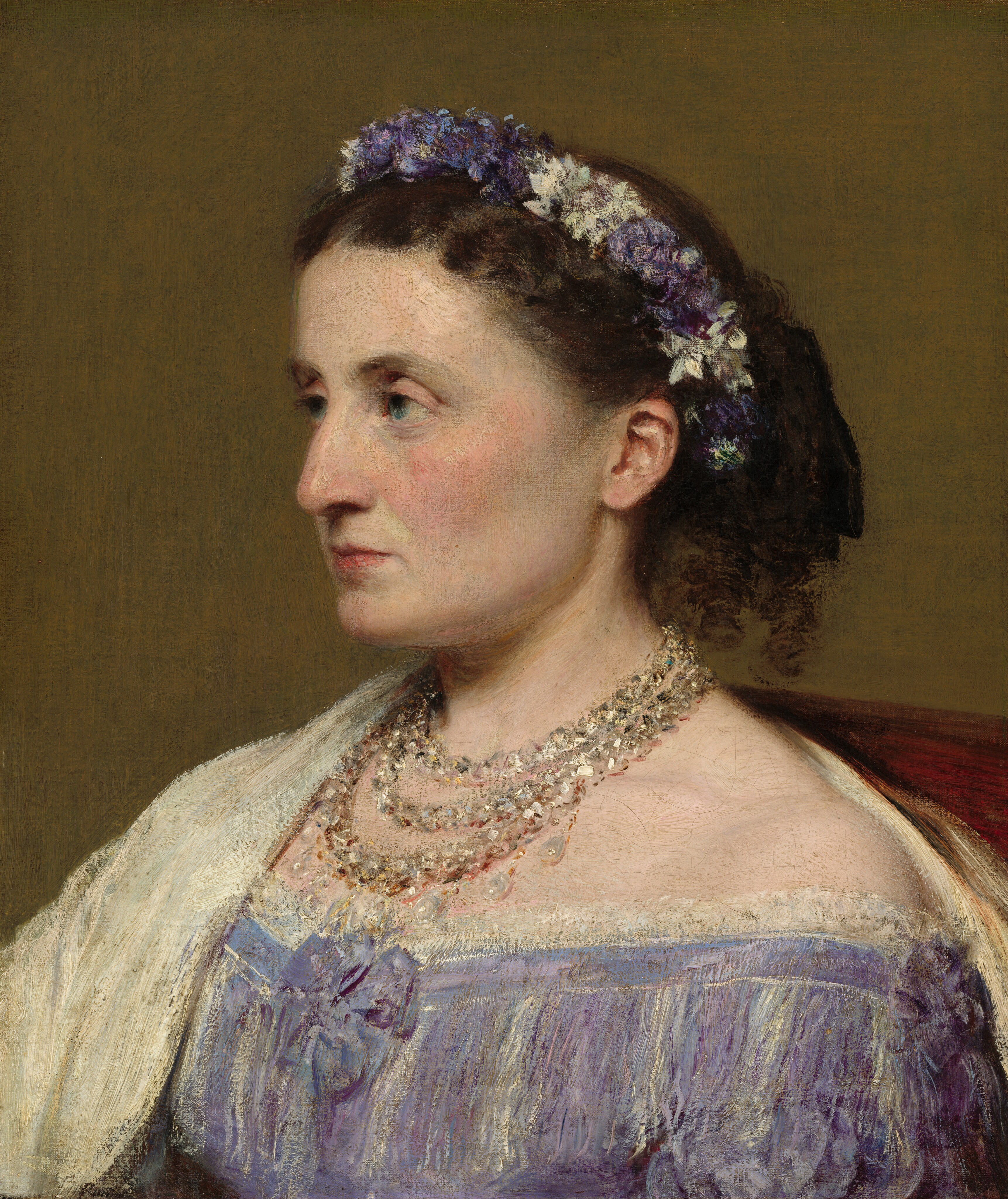 This painting is a detailed portrait featuring the upper body of a woman seen in side profile, facing the left. She has a fair complexion, with a curved nose, pale eyebrows, pink cheeks, high cheekbones, deep-set eyes, and thin pink lips in a neutral expression. Her curly, dark brown hair is styled behind her head and adorned with a wreath of purple and white flowers. She wears a lilac-purple off-the-shoulder dress with white fringe details and a bow in the front. A light-colored shawl or wrap drapes over her shoulders, and she wears multiple strands of pearl necklaces. She appears to sit in a dark red chair that is partially visible on the right, and the background is plain, muted brown.