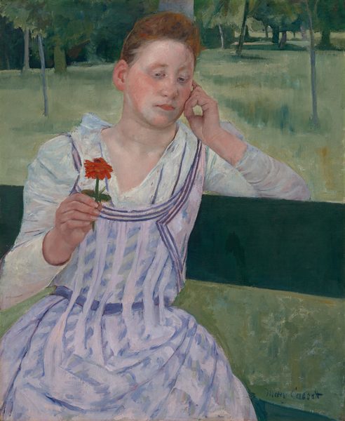 Shown from the knees up, a young woman with peachy skin and red hair sits facing us on a pine-green bench in this vertical painting. She sits with her body angled to our right, and her left elbow, on our right, hooks over the top slat of the bench. Her head tilts slightly to our right to rest on the curled fingers of that hand. In her other hand, she holds up a red flower on a short stem. Her hair is pulled up and back over a high forehead, and her cheeks are deeply flushed. Her pink lips are closed, and she looks down and to our left at the flower with gray eyes. She has an oval-shaped face and a hint of a double chin. Her white dress is painted with ice-blue shadows and folds. It has a deep V neck that reaches to an apron-like pinafore patterned with vertical bands of pale lilac that alternate with angled daubs of blue. Beyond her is a loosely painted landscape with grassy areas and clusters of tall trees painted in spring, emerald, and sea glass green. The edge of the grassy field nearly brushes the top edge of the canvas, and the trees are cropped by it. The artist signed the lower right, “Mary Cassatt.”