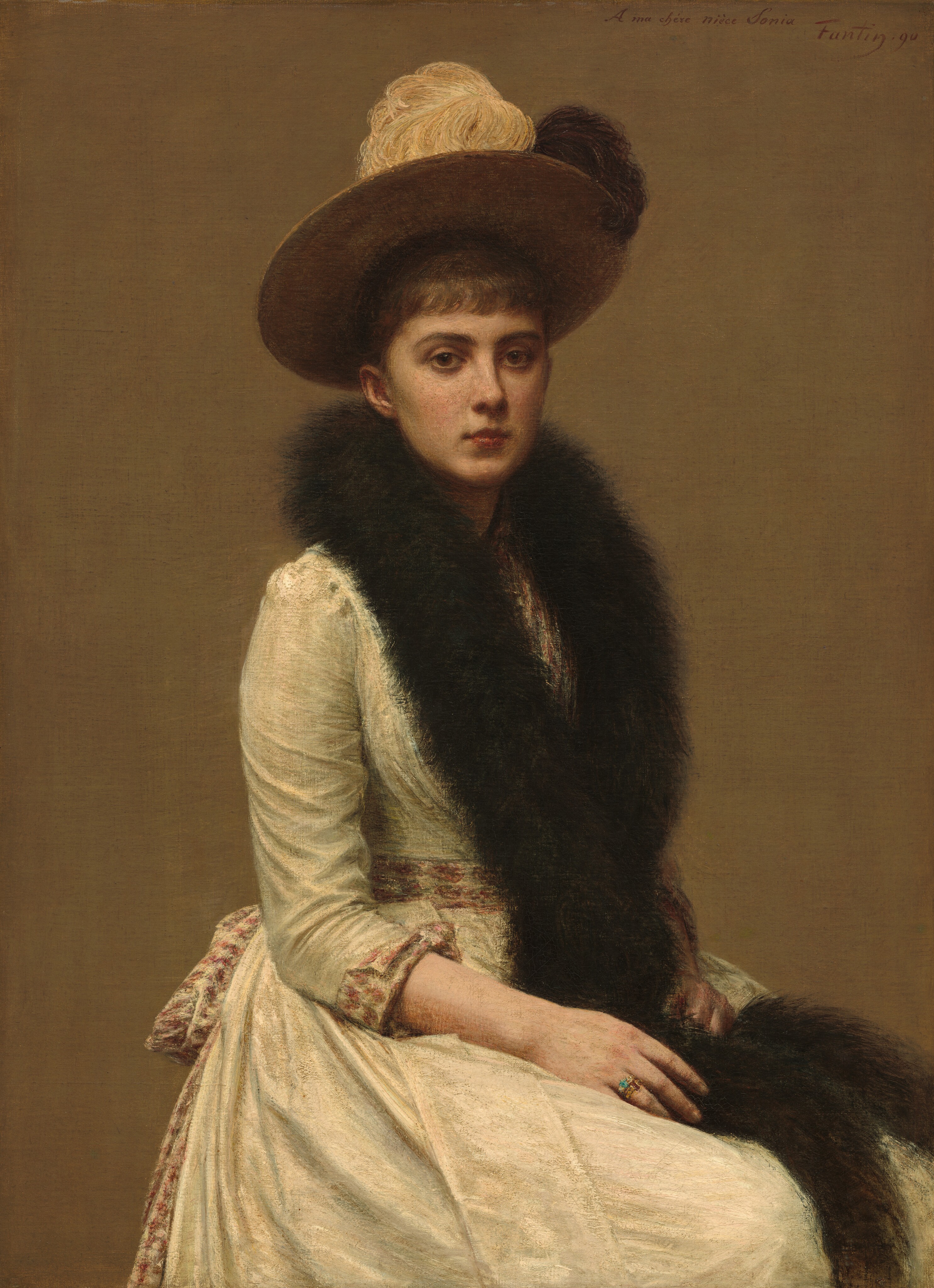 Shown from the knees up, a young pale-skinned woman sits looking at us in this vertical portrait painting. Her body is angled to our right, but she turns her face to gaze out with large brown eyes under dark brows. She has an upturned nose, and a wine-red, bow-shaped mouth in a narrow face. A fringe of brown bangs falls across her forehead, and her hair is pulled back under a wide-brimmed hat topped with a cream-white and dark brown feather. The cap is worn back on her head so only the cinnamon-brown underside of the brim is visible. A long black or very dark brown fur stole drapes around her neck, crosses at her waist, and falls to her lap. She wears an ivory-white gown with a muted burgundy pattern on the cuffs and matching sash that wraps her waist and ties in a bow at her back. Both hands rest in her lap. The closer hand lies alongside the stole, and she wears two gold rings on the third finger. One is set with a pale blue gem and the other with a dark red stone. Her other hand is a loose fist around the far end of the stole. She is set against a flat, camel-brown background. The artist signed and dated the upper right corner, “A ma chére nièce Sonia. Fantin. 90.”