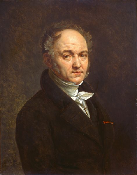 The image shows a man from the waist up. He is slightly turned to the side, with a serene expression. He has short, swept-back gray hair, sideburns, and visible lines on his face. The man is wearing a dark suit, white high-collared shirt, and a white cravat. A chain, possibly a watch fob, is visible on his suit. There is a red detail, possibly a ribbon or handkerchief, in his breast pocket. The background is plain dark brown.