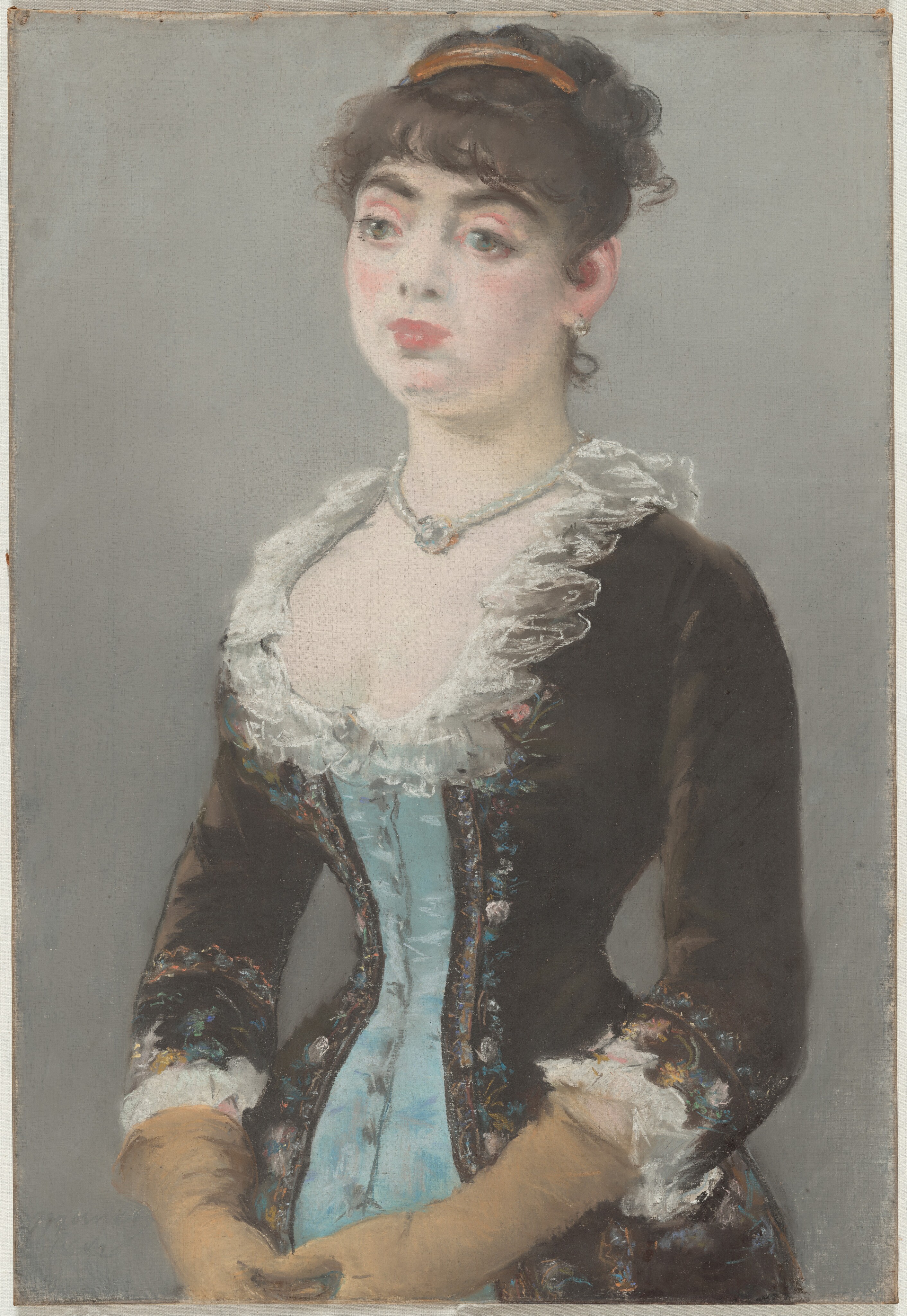 Shown from the hips up, a pale-skinned woman wearing a tightly corseted dress looks off to our left in this vertical pastel drawing. The portrait is loosely sketched with blended areas, giving it a soft look. The woman’s body is angled to our left, and her hands touch in front of her lower belly. She looks off with large, wide set, slightly bulging green eyes under dark brows. She has a round face, full pink lips, and the suggestion of a dimple in her chin. Her dark brown hair is pulled up under a gold band, and bangs curl across her forehead. A short, delicate ringlet curls up the nape of her neck. She wears a light-colored, round earring in the ear we see and a string of pearls, which may hold a round jewel that rests in the hollow of her throat. Her dress is powder blue down the front and coffee bean brown to either side and on the sleeves. The edges of the brown sections are lined with flowers drawn loosely in shades of pastel pink, blue, yellow, and green. The low, curving neckline is edged with white ruffles, and the waist is cinched to an almost startling degree. The sleeves come down just past her elbow, and the cuffs are also lined with white ruffles. Long, tan-colored gloves stretch back to the cuffs. The background is fog gray. The artist signed and dated the pastel faintly in the lower left corner, “Manet 1882.”
