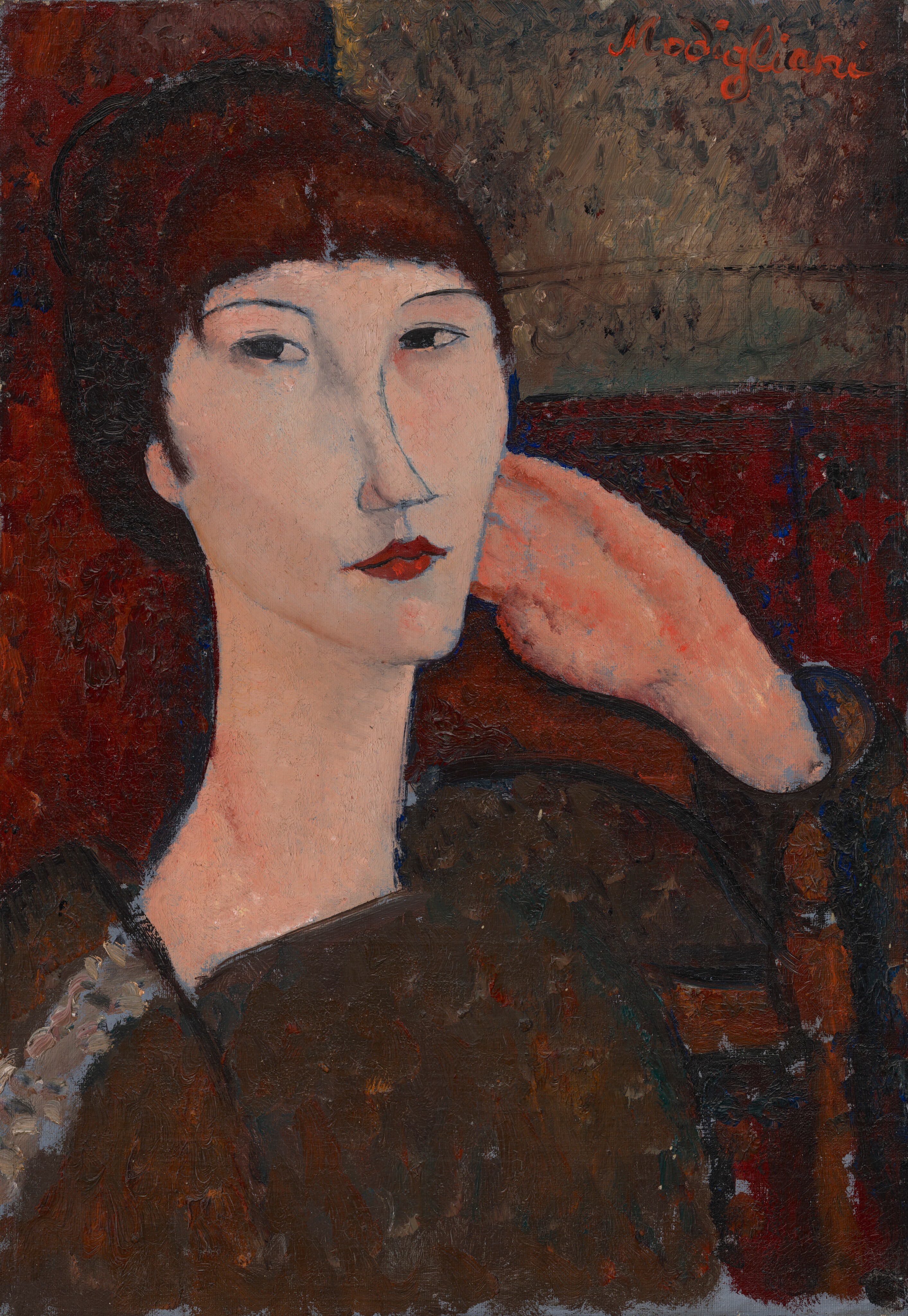 A woman with peachy skin and dark auburn hair is shown from her chest up sitting in a wooden chair in this vertical portrait painting. Her body is angled to our right, and her left arm, farther from us, rests on the back of the chair so the fingers of that hand rest alongside her left cheek. Her face is angled to our right but her dark eyes cut back to look at or toward us from the corners of her almond-shaped eyes. She has thin, gently arched brows, a long, sloping nose, and her burgundy-red lips are closed. Her features are outlined with delicate, dark gray or black lines. Bangs sweep across her forehead, and her hair looks like it might be pulled up and back. Her rust-brown garment has a square neckline, and a lighter strip of smoke gray and sky blue runs along her right shoulder, closer to us. She sits in front of an indistinctly patterned wine-red wall, with a lighter, tan-colored rectangle in the upper right corner. The background, her clothing, and the hand by her face are painted with areas of mottled color but the skin of her face seems smooth. The artist signed the painting with bright red letters at the upper right, “Modigliani.”