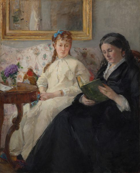 An older woman wearing a long black dress sits reading a book in front of a younger woman wearing cream white, who sits on a couch just beyond, in this vertical portrait painting. The scene is loosely painted with some visible brushstrokes, especially in the furniture, clothing, and room. Both women have pale skin and wear dresses with long sleeves and skirts. To our right, the older woman sits in a chair with a curved back, which runs parallel to the right edge of the canvas. The full skirt of her black dress angles down across the picture, almost spanning the bottom edge of the composition. Her gray hair is pulled back and covered by a black cloth or veil, which falls to her shoulder and blends with her dress. The collar is lined with a white ruffle, and white lace cuffs encircle her wrists. Her face is painted with rosy tones. She looks down at her book with dark eyes, and her rose-pink lips are closed. She wears a gold ring with a light stone on her left ring finger, and the book she holds has a moss-green cover. Her skirt overlaps the younger woman sitting nearby on the couch, which runs along the back wall of the room. The young woman’s knees are angled to our left but she looks slightly back to our right, to gaze down and into the distance with brown eyes. Her skin is paler, but she has lightly flushed cheeks and her petal-pink lips are also closed. An azure-blue bow is tied in her chestnut-brown hair. A few tendrils fall down her back and over one shoulder. Her dress falls loosely over her torso and legs. She wears a gold ring on her left hand and a gold bracelet on that wrist. She fingers a piece of lapis-blue fabric in one hand. The couch behind her has a pattern with stylized flowers or leaves floating within rose-pink ribbons that hang in swags, against a white background. Pillows patterned with ruby red, sky blue, and fawn brown peek out from behind the young woman. A round or oval wooden table with curving legs to our left holds a glass vase with a violet-purple flower, perhaps a hydrangea. A slip of paper or envelope sits on the table next to the vase. The wall behind the couch is fog gray, and the bottom edge of a gold frame hangs behind the young woman.