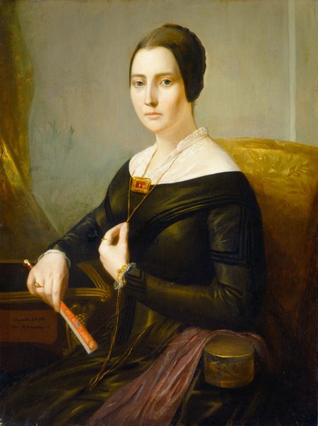 The image shows a woman seated in a chair. She has dark brown hair in a neat updo, soft facial features, and light skin. She wears a black gown with a white lace collar, a gold necklace with a red pendant, a bracelet, and rings. She holds a red closed fan in her right hand. The background is a light-colored wall, and she sits on a chair with golden upholstery. A small round box is placed on the chair's armrest to her right.