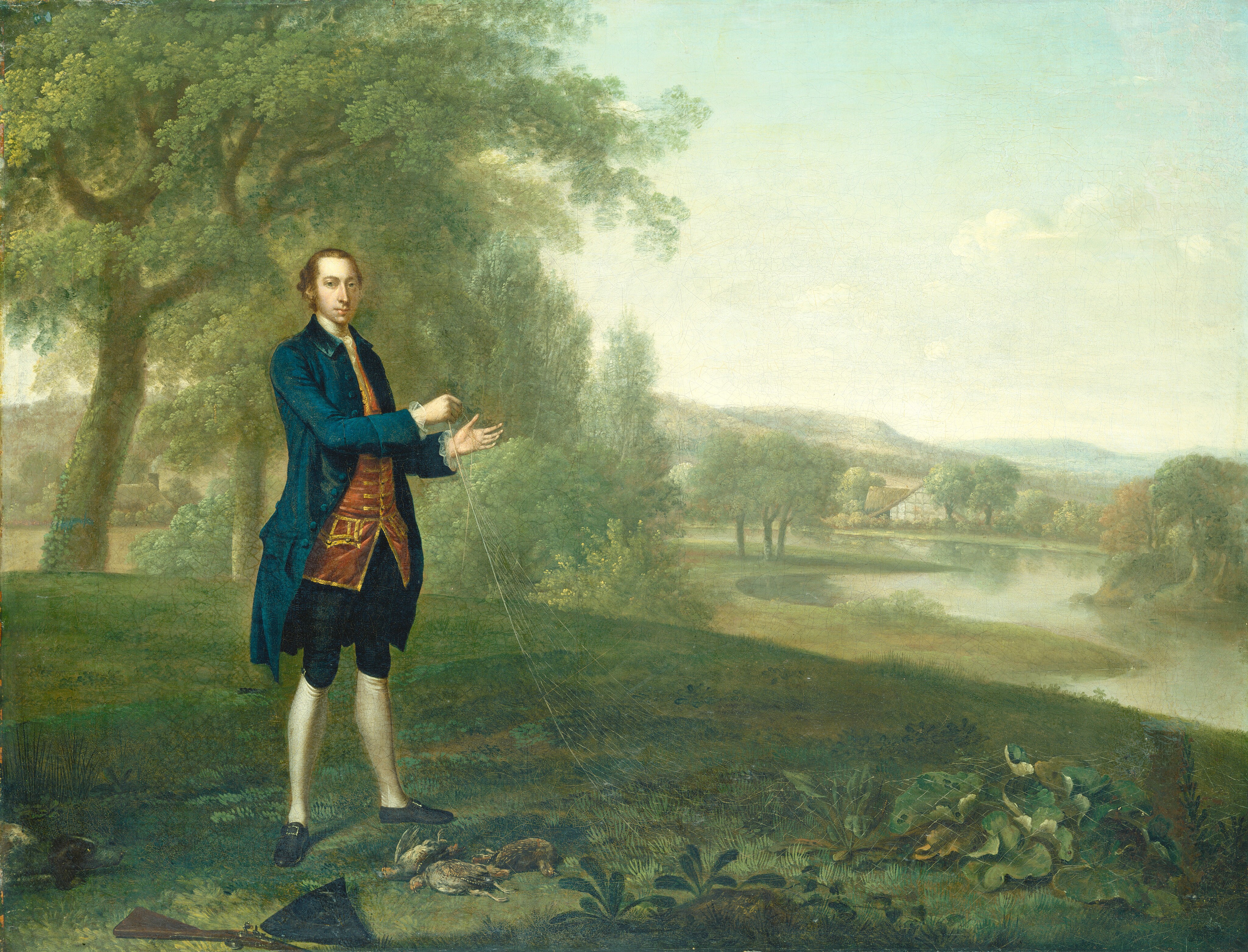 The painting shows a man standing in a lush green landscape. He is elegantly dressed in an 18th-century outfit, consisting of a dark blue coat, a red waistcoat and knee-length breeches, with white stockings and black shoes. He appears to be holding strings or cords. To his right, guns and game birds are placed on the grass. A river can be seen flowing gently in the mid-level of the painting. Close by, there are tall trees with dense foliage, painted with detailed leaves. In the distance across the river, a house is visible nestled amongst more trees, and the landscape extends into softly rolling hills. The sky is pale blue with light clouds, complementing the serene, verdant palette of greens and browns that dominate the scene.