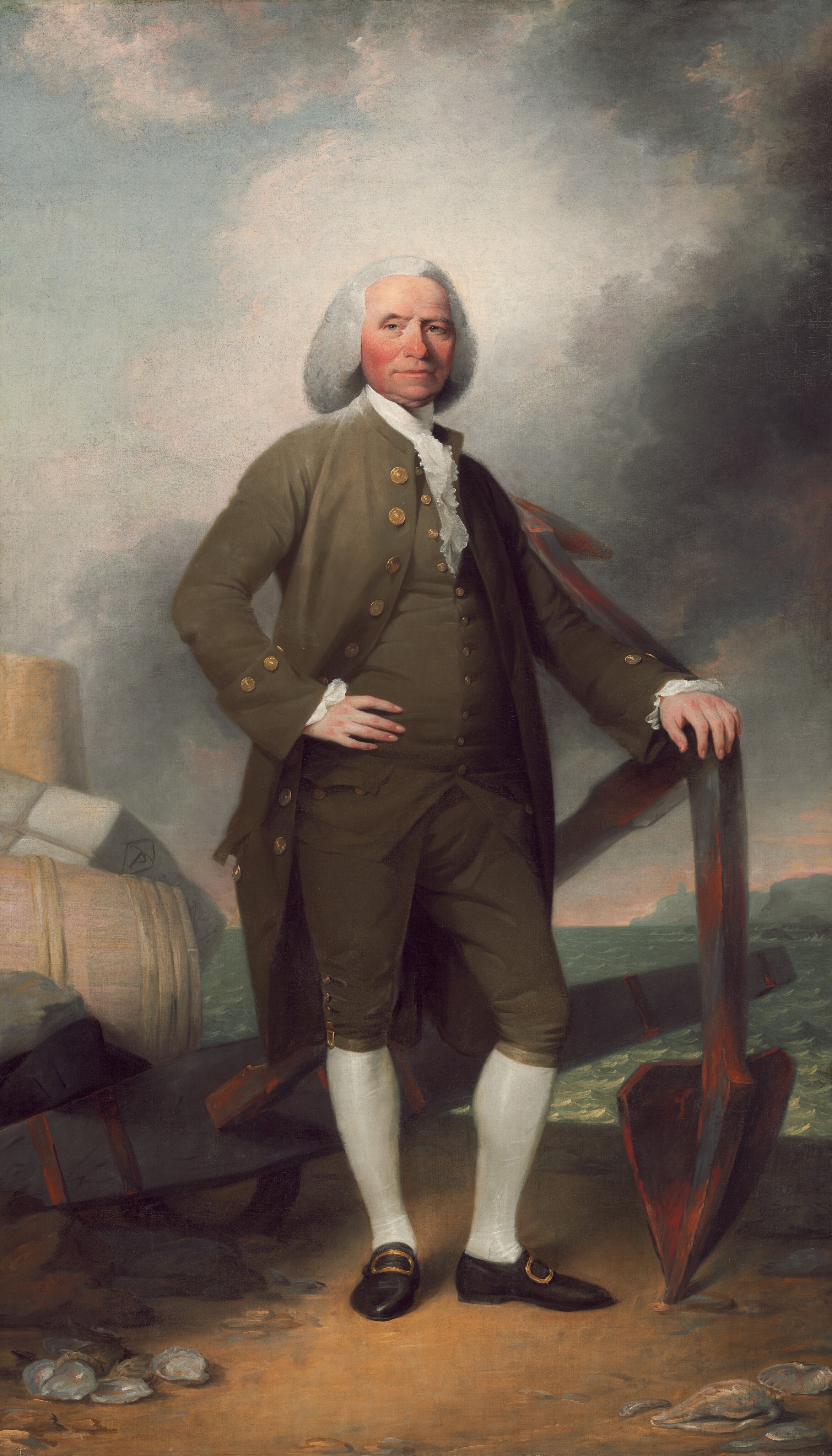 This painting shows a man standing confidently with one hand on his hip and the other resting on a large, rusty anchor. His body is slightly turned to the right while his head is turned towards us, and one of his knees is slightly bent. The man has fair skin with rosy cheeks and a neutral expression. His hair is white and styled in faint curls that fall down around his chin and the tops of his shoulders. He wears an olive-green coat with brass buttons, a matching waistcoat over a white cravat, knee-length, olive-green breeches, white stockings, and black shoes with brass buckles. He stands on the edge of a sandy shore, and to the left are a pile of barrels and crates. The background includes a cloudy sky and a glimpse of the sea with waves and distant cliffs visible in the distance.