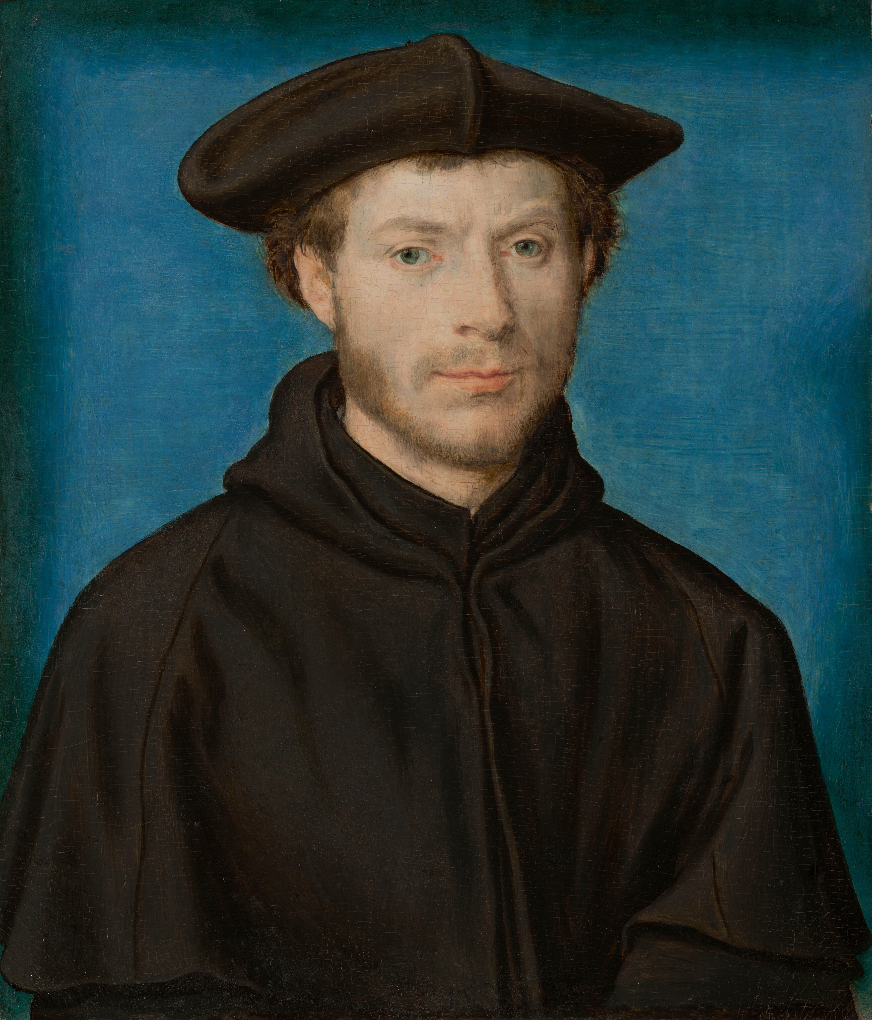 This painting shows a man from the waist up. He is facing forward and gazes out at us. He has fair skin, light brown hair, a thin mustache and beard, and light green eyes. His expression is mostly neutral, although his peach lips are slightly turned up at the edges. He is wearing a black garment with a high collar and a hood and a black flat cap on his head. The background is plain blue, fading to black around the edges of the painting.
