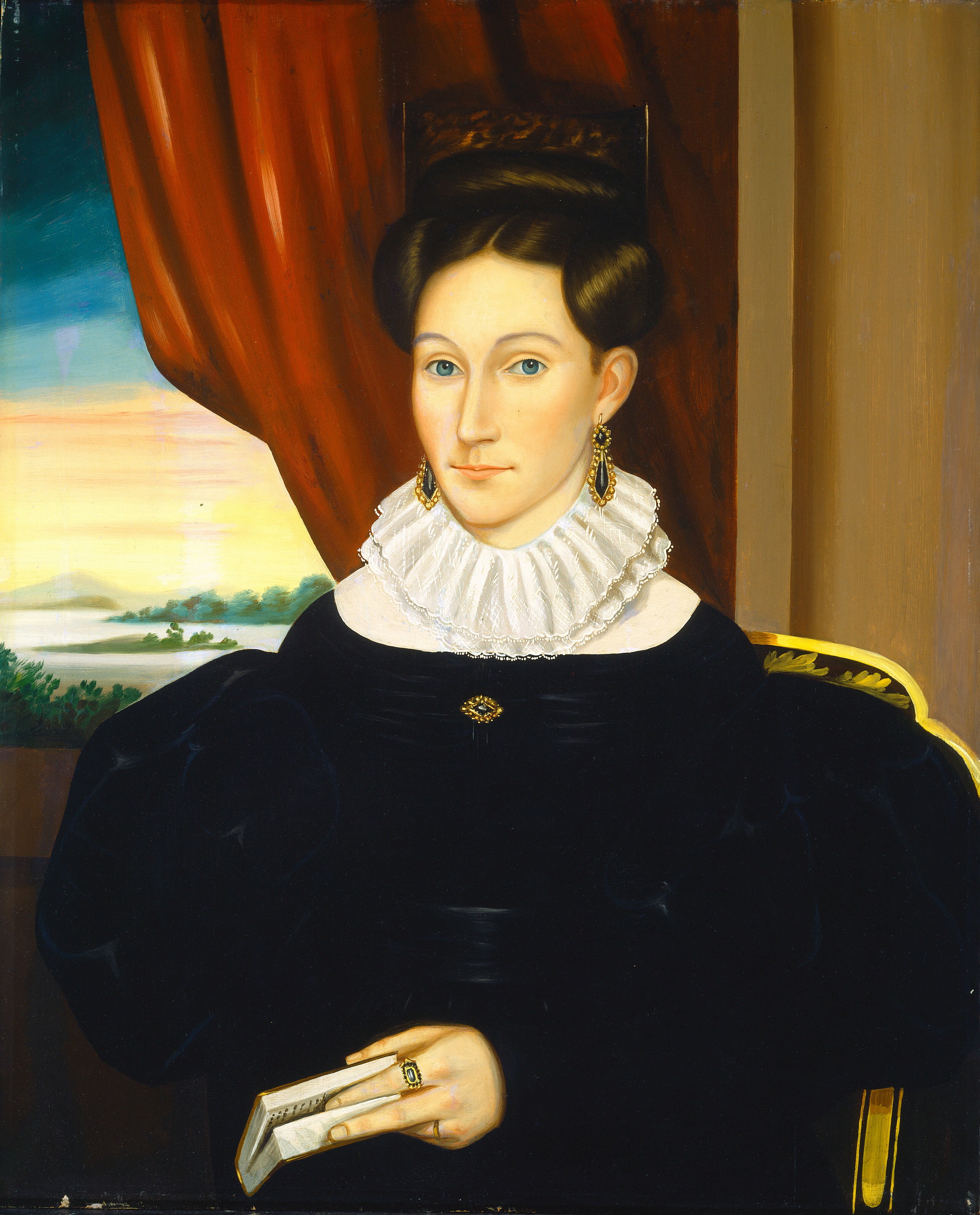 The image shows a half-length portrait of a woman seated in a chair. She is facing slightly to her right, with her gaze directed towards the viewer. She has fair skin, blue eyes, and a solemn expression. Her hair is dark brown and styled in an elaborate updo with large, smooth rolls on either side of her head. She wears a black dress with voluminous sleeves and a high white ruffled collar. She has a brooch around her neck, gold earrings, and a ring with a green stone on her left hand, which is holding a book. The setting features a dark red curtain behind her, partially pulled back to reveal a countryside landscape through a window. A portion of the chair with a curved golden edge is also visible to her left.