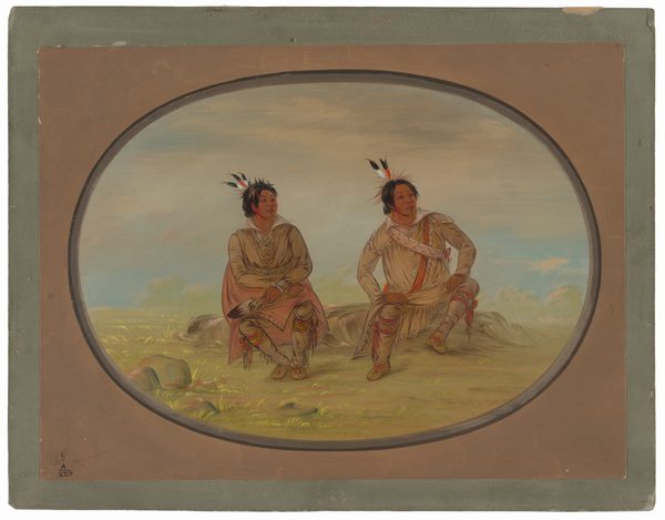 The image includes two seated figures, both shown from the knees up. Each person is seated with their legs bent and feet resting on the ground. The individual on the left has dark hair with feathers and wears a long garment with a pattern across the chest and over the shoulders. Their garment reaches the knees, revealing moccasin-style footwear. This person holds an object in their hand, which appears to be a feather fan or similar item. The figure on the right also has dark hair with feathers and wears a long-sleeve top with a decorative sash, knee-length leggings, and moccasins. The background shows an open landscape with green plains, rocks, and a cloudy sky, suggesting an outdoor setting. The image is enclosed within an oval frame.