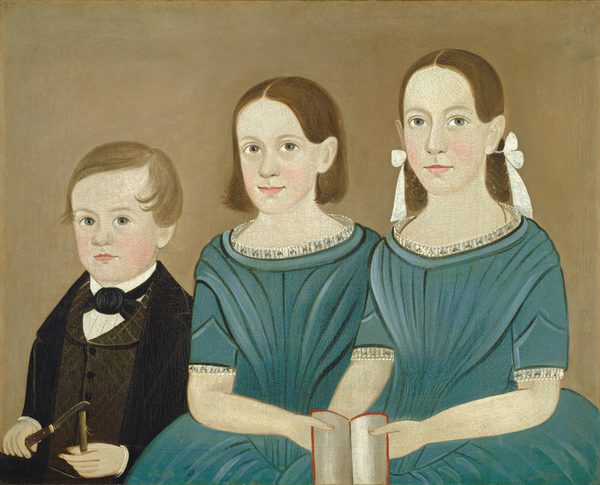 The image shows three children in a seated position from the waist up. The child on the left is a young boy with short, light brown hair. He wears a black suit with a bow tie and a white shirt and holds a small object. The two children on the right are young girls with shoulder-length brown hair and large white bows. They wear matching blue dresses with silver embellishments. The girl in the middle holds a book. The background is plain beige.