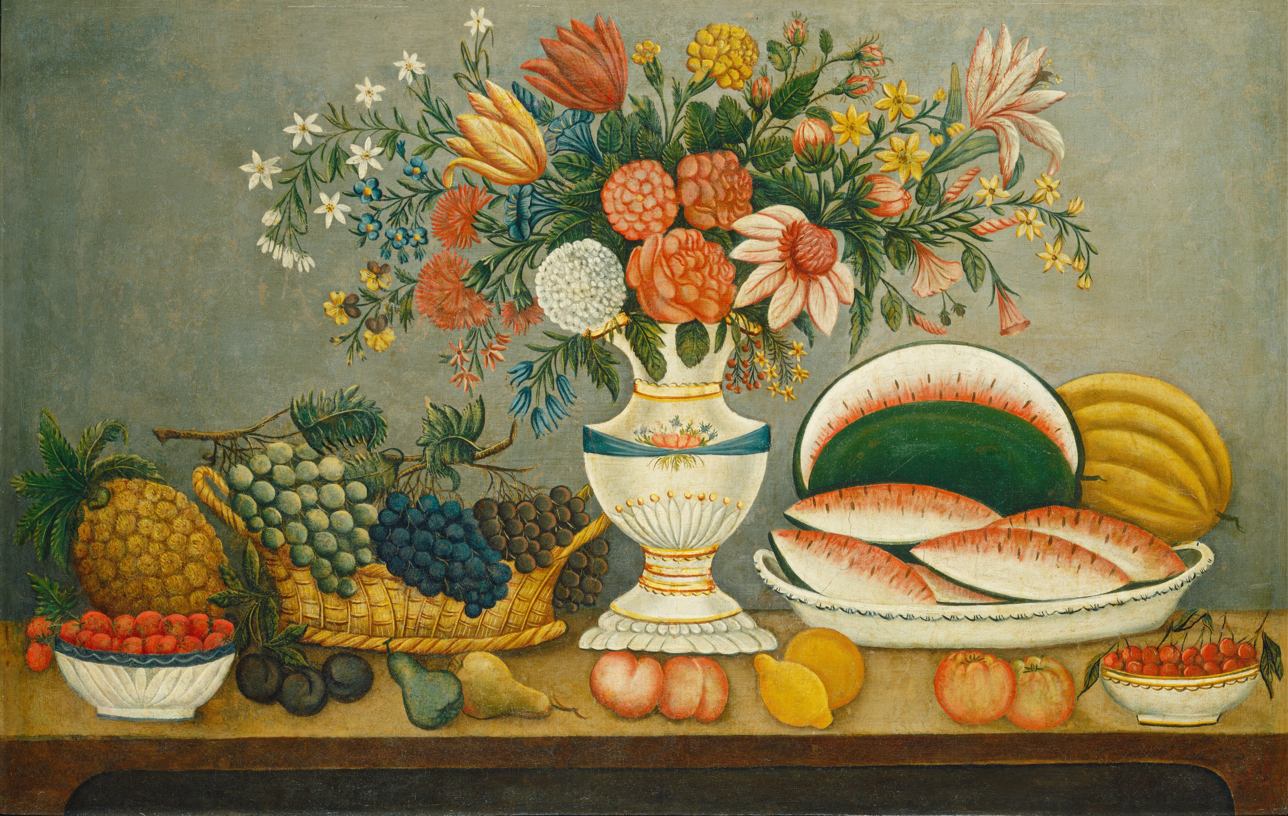 In this painting, an ornate vase filled with colorful flowers rests on top of a long brown table. A variety of fruit is spread around the vase. On the left side of the painting, there are red and green grapes in a wicker basket, a pineapple, red berries in a small white bowl, three small plums, and two pears. On the right side is a platter with watermelon slices and a yellow gourd or pumpkin, two yellow lemons, two peaches, two apples, and red cherries in a bowl. The flowers in the vase are a variety of colors including red, pale pink, white, yellow, and blue, with green flowers and stems between them. Behind the table is a light green wall.