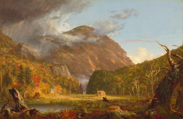 We look across a wide, low clearing at a mountain rising beyond rocky hills, which are mostly covered in pines and blazing red and orange autumn trees, in this horizontal landscape painting. The green grass of the clearing is littered with dead trees and stumps. Beyond a narrow opening between the hills, almost at the center of the composition, a massive mountain towers against the powder-blue sky, nearly reaching the top edge of the painting. The mountain’s jagged face is mottled with warm tones of cinnamon, rust, and caramel brown. The scene is bathed in warm light coming from our right but pewter-gray storm clouds move in from the upper left to encircle the mountain peak. Closest to us, a line of broken tree trunks spans the width of the painting, mixed with scarlet-red, marigold-orange, and green vegetation and reeds. A silvery pond lies just beyond them to our left. Tiny in scale within the landscape, a horse and rider gallop along a wide dirt road emerging from the lower right toward a white house nestled at the foot of the hill to our left. The rider wears a knee-length, denim-blue tunic and red leggings. Upon closer inspection, two children and a white dog become visible, walking toward the rider. A straw-yellow structure sits against the hill to our right. A horse-drawn carriage moves away from the house in the far distance, where the road curves toward the notch. The artist signed and dated the painting as if he wrote directly onto a broken trunk in the lower left corner of the painting: “T. Cole 1839.”