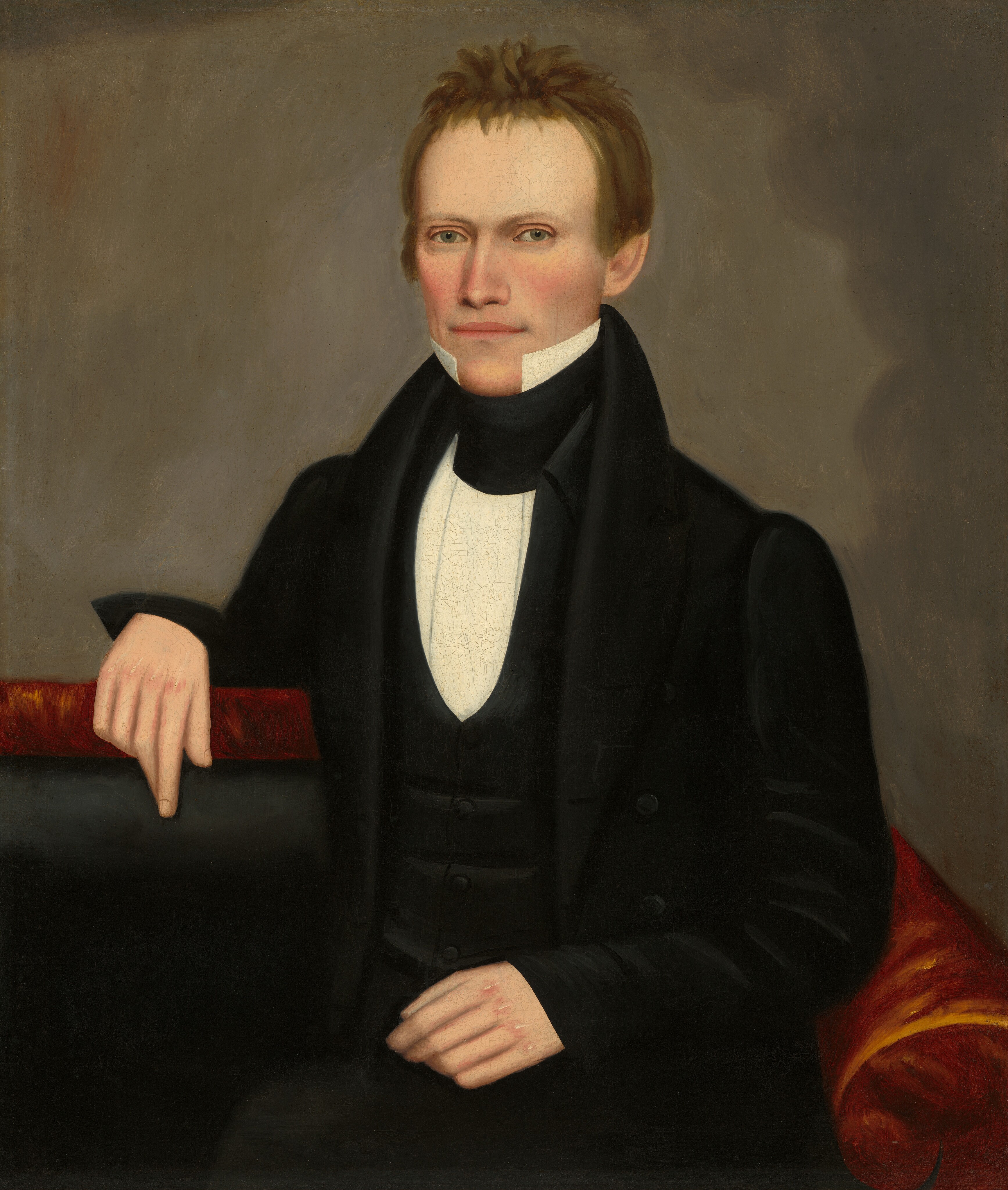 The image depicts a man shown from the waist up. He is facing slightly to the right, with his left arm draped over the back of a red chair and his right hand resting on his lap. He has a fair complexion, clean-shaven face, prominent cheekbones, and deep-set eyes. His short light brown hair is slightly tousled. The man is wearing a black coat, a black waistcoat, a white high-collared shirt, and a black cravat. The background is a muted dark brown color.