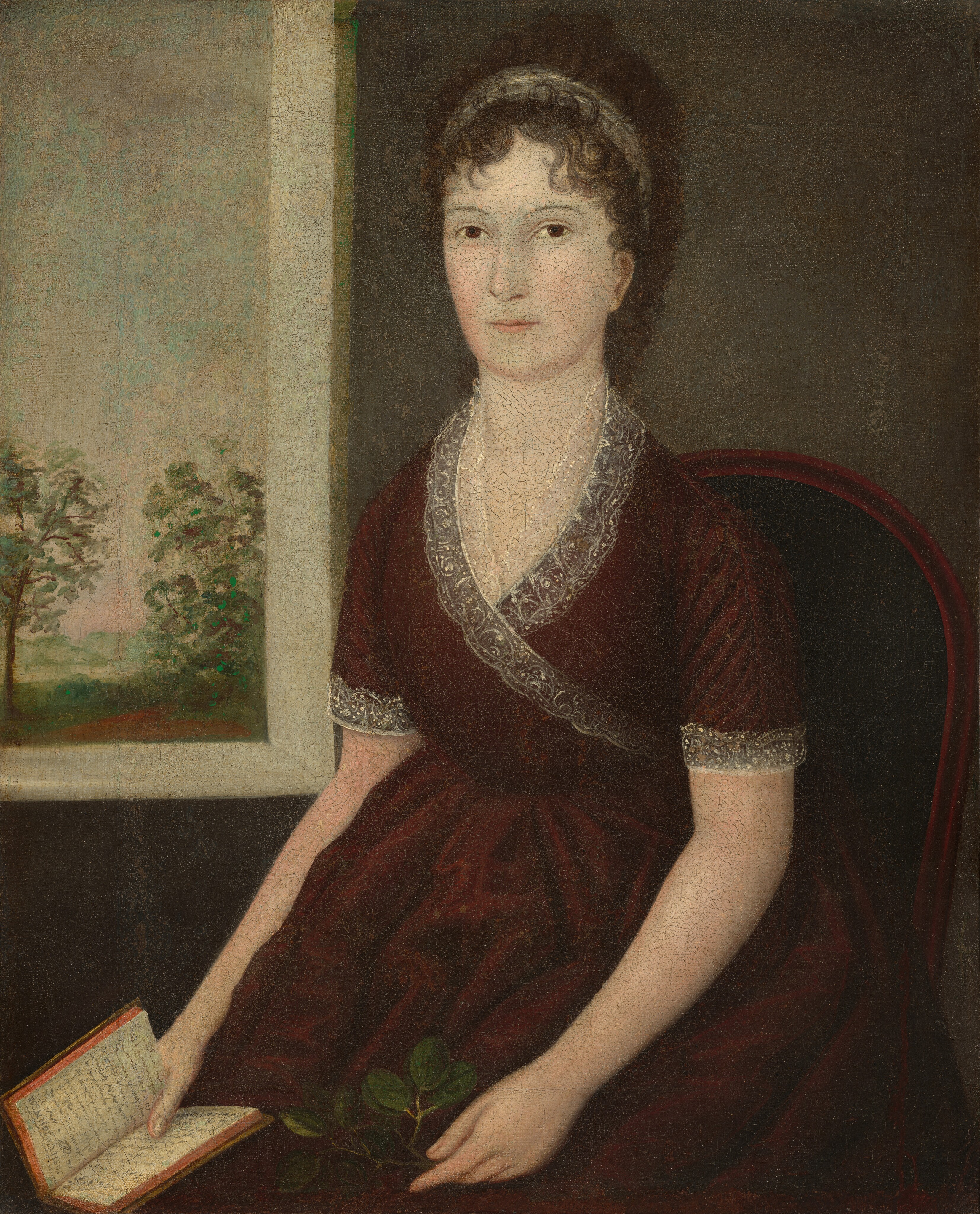 This painting shows a seated woman depicted from the knees up. She is sitting in a dark reddish-brown chair with a dark green cushion, her body turned slightly to the left, facing the viewer. She has both arms resting on her lap, her left hand holding a small twig with round green leaves and her right hand holding a small book open. She has light skin, dark brown eyes, thin eyebrows, thin pink lips set in a neutral expression, and dark curly hair styled away from her face and adorned with a white headband. She wears a dark red dress with lace detailing around the neckline and the cuffs of the short sleeves. The background features a dark gray wall on the right, with a white window on the left providing a view of an outdoor scene with leafy green trees and a sky that is a gradient of light gray and light blue, with light pink at the horizon.