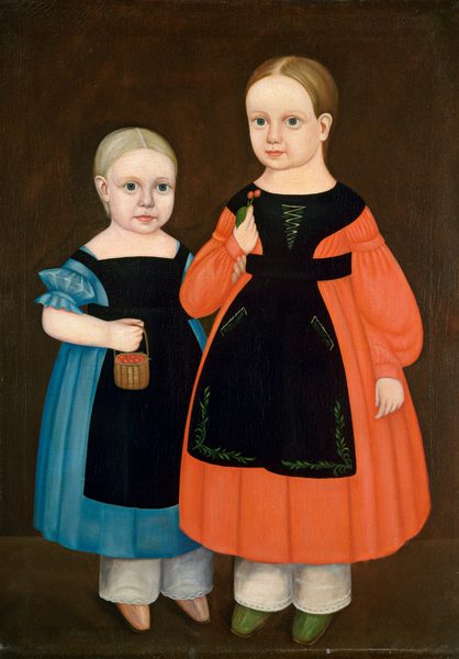The image shows two young children depicted in a full-body portrait, standing side by side. The child on the left has light-colored hair pulled back tightly, large blue eyes, and a fair complexion. This child is wearing a blue dress with puffed sleeves, a black apron, and white undergarments with visible lace at the hem. They are holding a small woven basket filled with red berries or similar objects in their right hand. The child on the right also has light brown hair, parted neatly in the middle, large blue eyes, and a fair complexion. This child is dressed in a bright orange dress with long sleeves, a black apron adorned with green embroideries, and white undergarments visible at the bottom. They are holding two red cherries with attached green leaves. The background is a plain, dark brown canvas which complements the vibrant colors of the children's clothing.