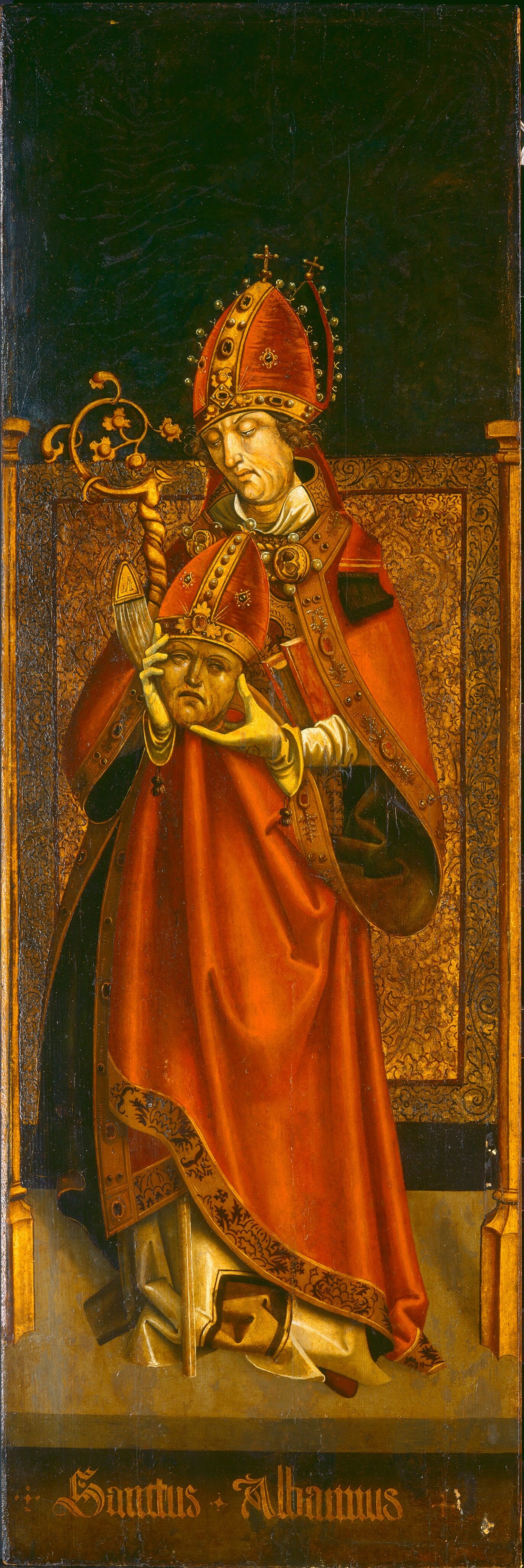 The image depicts a figure identified as "Sanctus Albanus." The individual is standing upright and adorned in ecclesiastical attire, including a bishop's mitre with jewels and a cross at the top, a red robe with gold trimmings, and a white vestment peeking out. The figure's face is serene, framed by curly brown hair. One hand holds a crosier, while the other cradles a mitred head. The background is dark, contrasting with the opulent gold and red attire. The intricate design work is visible on the clothing and a golden panel behind the figure. The base of the panel bears the text "Sanctus Albanus" in decorative lettering.