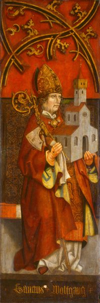 The image depicts a person standing upright, facing forward and slightly to the side. The individual has a solemn expression, with defined facial features and light-colored hair. They wear ecclesiastical attire, including a red outer garment with wide sleeves and a white inner robe. The person wears an elaborately designed mitre with intricate patterns. They hold a bishop's crozier in one hand and a model of a church in the other. An ornate large medallion hangs around their neck, suggesting high status. The background shows rich golden ornamental patterns against a deep red backdrop.