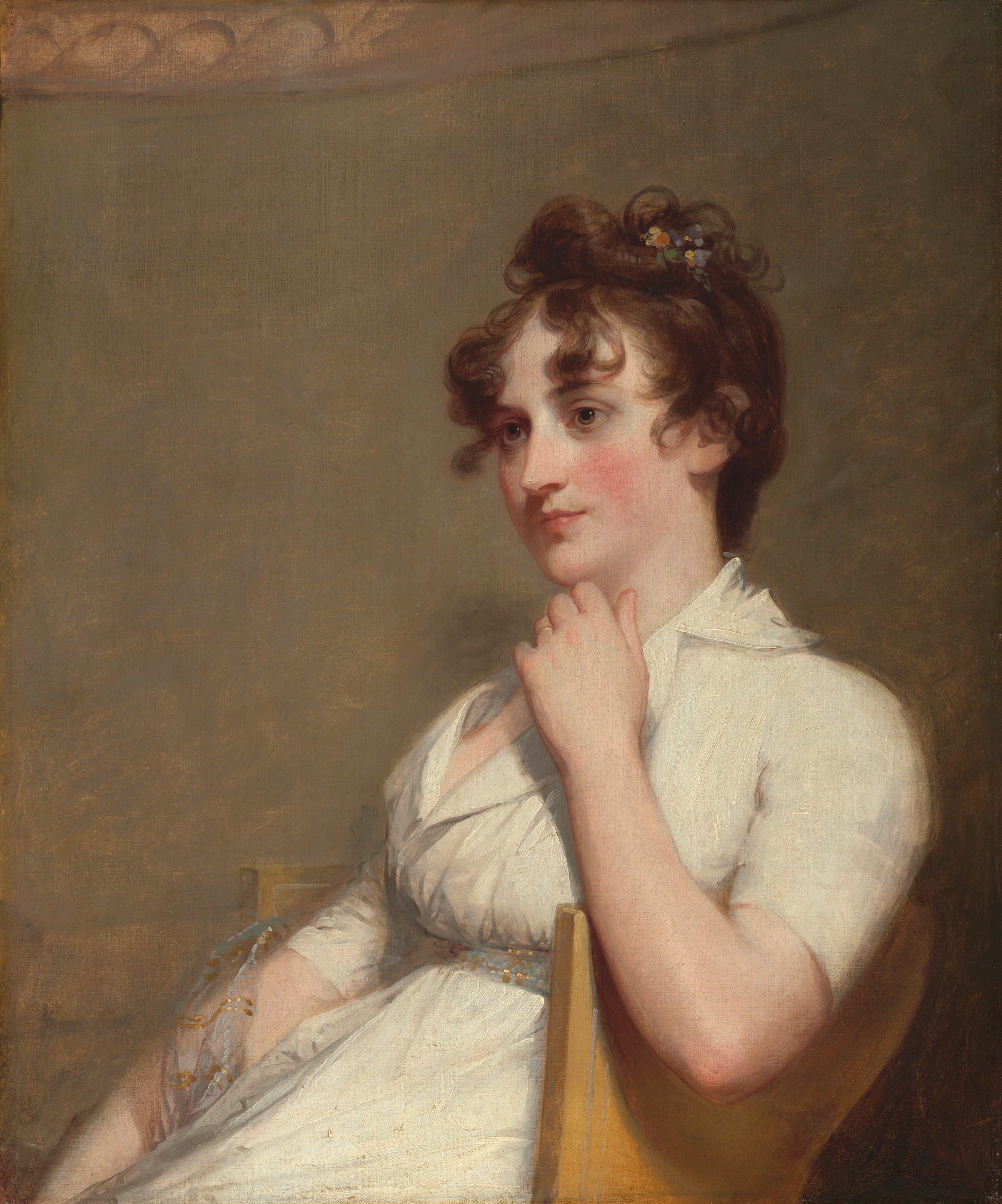 This painting is a portrait of a seated woman looking off to the left with a composed expression. She has light skin with rosy cheeks, brown eyes, and slightly upturned pink lips. Her curly brown hair is styled on top of her head with a small floral clip or decoration. She is wearing a white dress with short sleeves and a low neckline with a wide collar. Around her waist is a thin belt with gold and light-blue designs, and a translucent cloth with a similar pattern is draped over her right forearm. Her other arm rests over the edge of the tan chair she sits in, and she brings her hand up slightly below her chin. A thin gold band is visible on one of her fingers. The background is a plain, muted brown color, with a curved tan section at the very top.