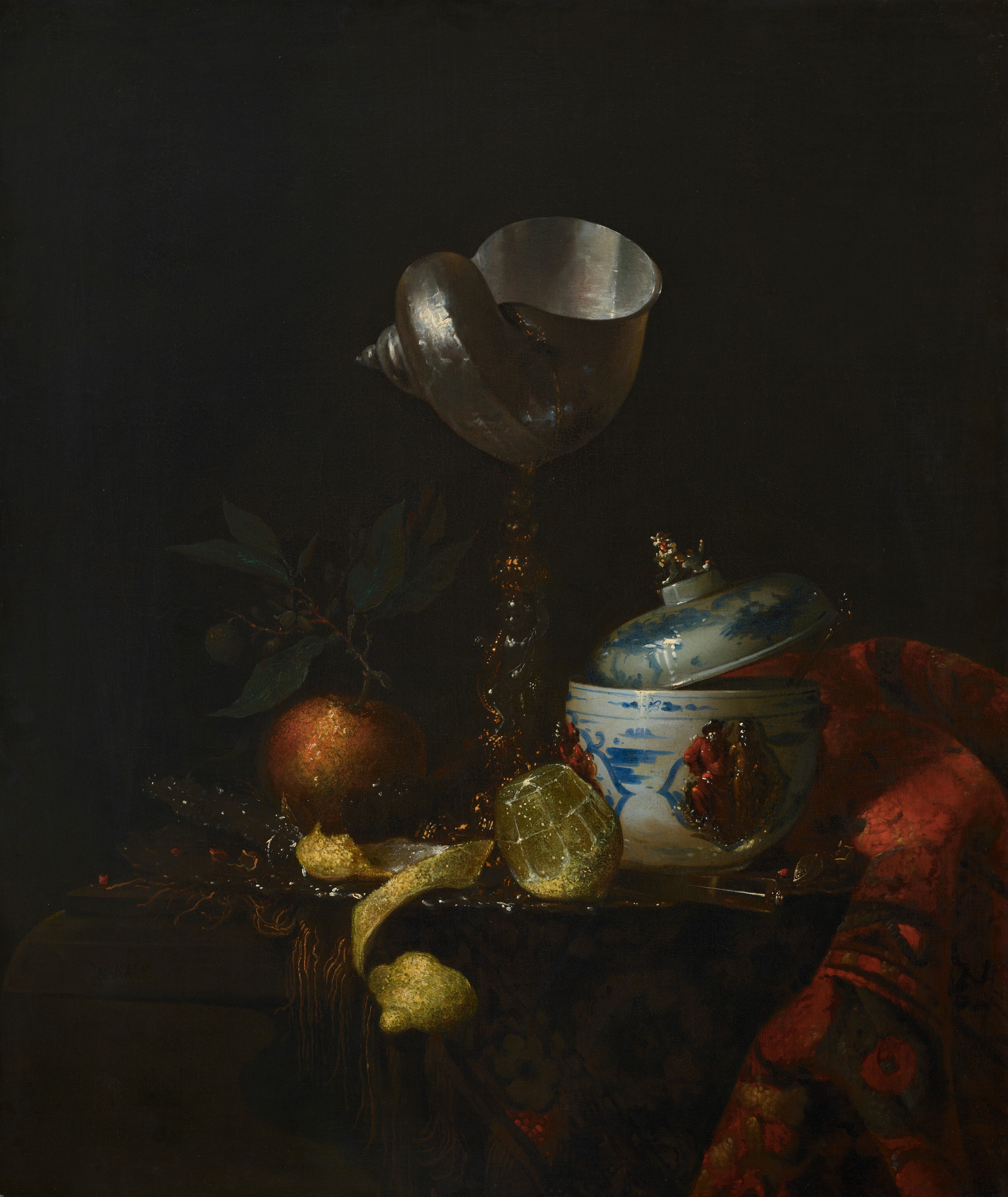 A tall goblet made with a spiraling nautilus shell on a gold and silver stem, a blue and white lidded, porcelain bowl, an orange, a cut lemon, and a bunched up, woven rug are gathered on a tabletop in front of a black background in this vertical still life painting. Light falling on the objects from our left contrasts sharply with the deep shadow beyond, which makes some details difficult to make out. The objects might be gathered on a silver tray. At the center of the composition, the flagon is made with a shimmering, ivory-colored nautilus shell with its wide opening facing up. The stem is made of gold bands coiling around a silver figurine, which is nearly lost in shadow. To our right, a white porcelain bowl has a rounded bottom and tall sides. It is painted with blue designs around a raised relief showing two people in crimson-red and harvest-gold garments. A utensil sits in the bowl to our right, and the lid, which has a finial in the shape of an animal, rests angled upward, partially in the bowl and partially on the stem of that utensil. A translucent, cylindrical knife handle protrudes toward us, angled to our right, in front of the bowl. A rug with a geometric pattern of rust red, tan, and pine green is bunched next to the bowl, along the right side of the painting. To our left of the nautilus cup, an orange with a stem and green leaves sits next to a bright, glistening lemon at the foot of the cup. Between the two pieces of fruit, the vivid yellow lemon rind has been peeled into a long, curling strip that curves off the front edge of the table.