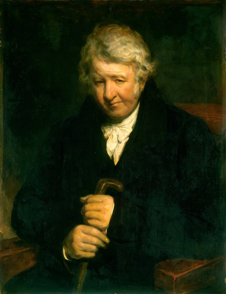 The image shows an older man sitting in a chair. He has a fair complexion, a round face, deep-set eyes, and white curly hair. He is dressed in dark, formal clothing and is leaning slightly forward with his hands resting on wooden armrests. One hand holds a wooden cane while the other covers it. The background is dark and unobtrusive.
