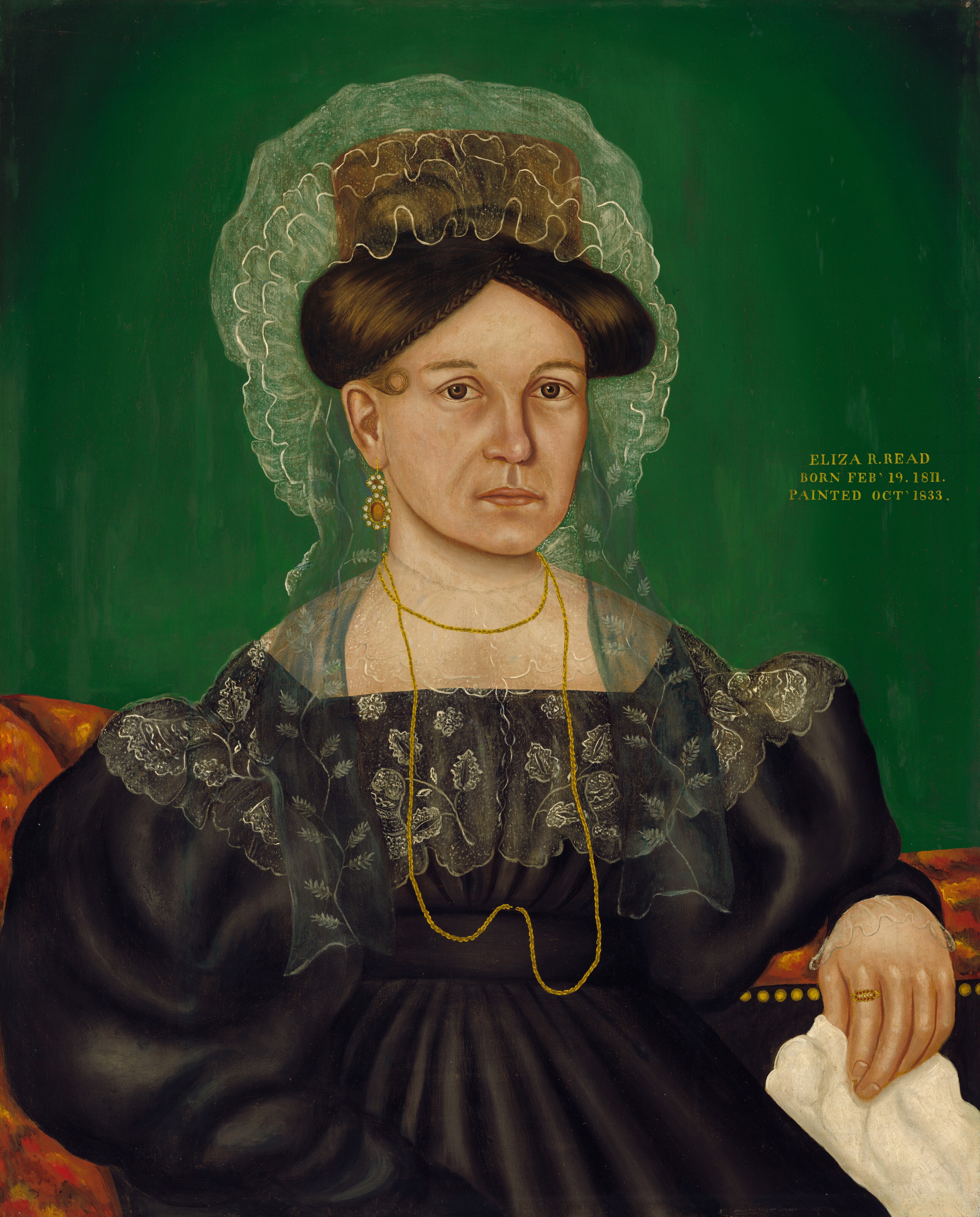 The image depicts a woman from the waist up. She is seated, facing slightly to her right but looking directly at the viewer. Her facial features are characterized by a solemn expression, with prominent cheekbones, light skin, and dark eyes. Her brown hair is styled tightly on top of her head, adorned with a delicate lace headpiece. The woman is dressed in a formal, black gown with voluminous sleeves, accented by a sheer, lace shawl with intricate patterns draped over her shoulders. She wears a long, fine gold chain around her neck, and a gold earring in her ear. In her right hand, she holds a white handkerchief. Behind her is a vibrant green background, with gold lettering to her left reading "ELIZA R. READ BORN FEB' 19. 1811. PAINTED OCT' 1833." She is sitting on an upholstered piece of furniture with a patterned orange design.