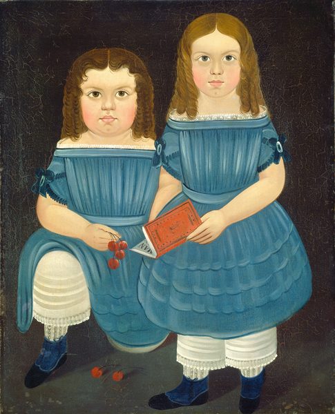 The image shows two young children standing together. The child on the left has fuller cheeks and tightly curled brown hair, while the child on the right has longer, less curly hair. Both are dressed in blue dresses with white lace-trimmed sleeves and white pantaloons with lace. They wear blue shoes. The child on the left is holding red cherries, some of which are on the ground, and the child on the right holds a red book with a partially visible white page. The background is a simple, dark tone.