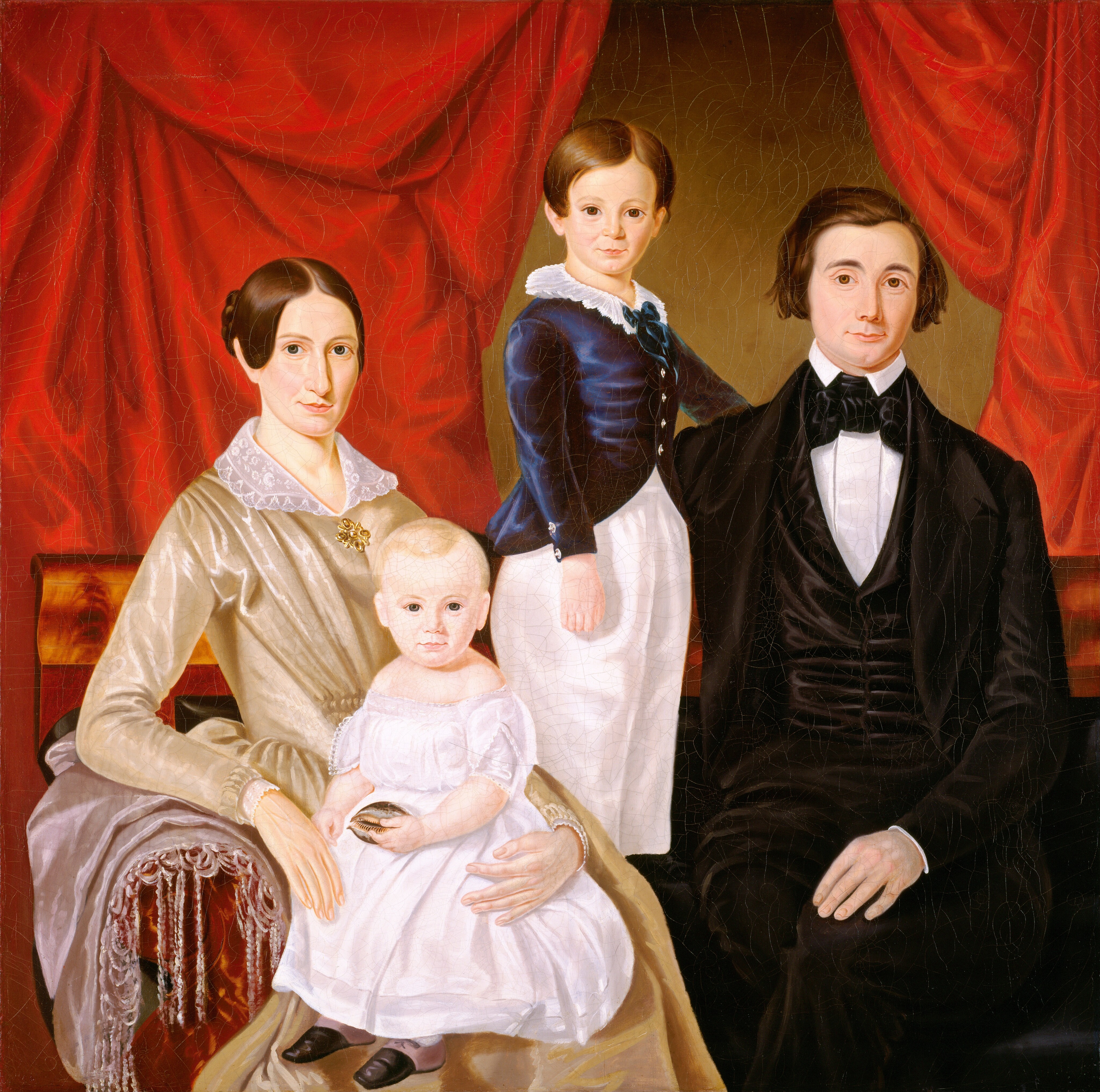 The image shows four individuals posing for a portrait. On the left is a woman in a light-colored dress with lace details and a gold brooch. She has dark hair styled in a bun and distinctive facial features. She is holding a young child in a white dress. Next to them is an older child wearing a blue jacket with a white collar. On the far right is a man in a dark suit with a black bow tie. The group is against a deep-red curtain.