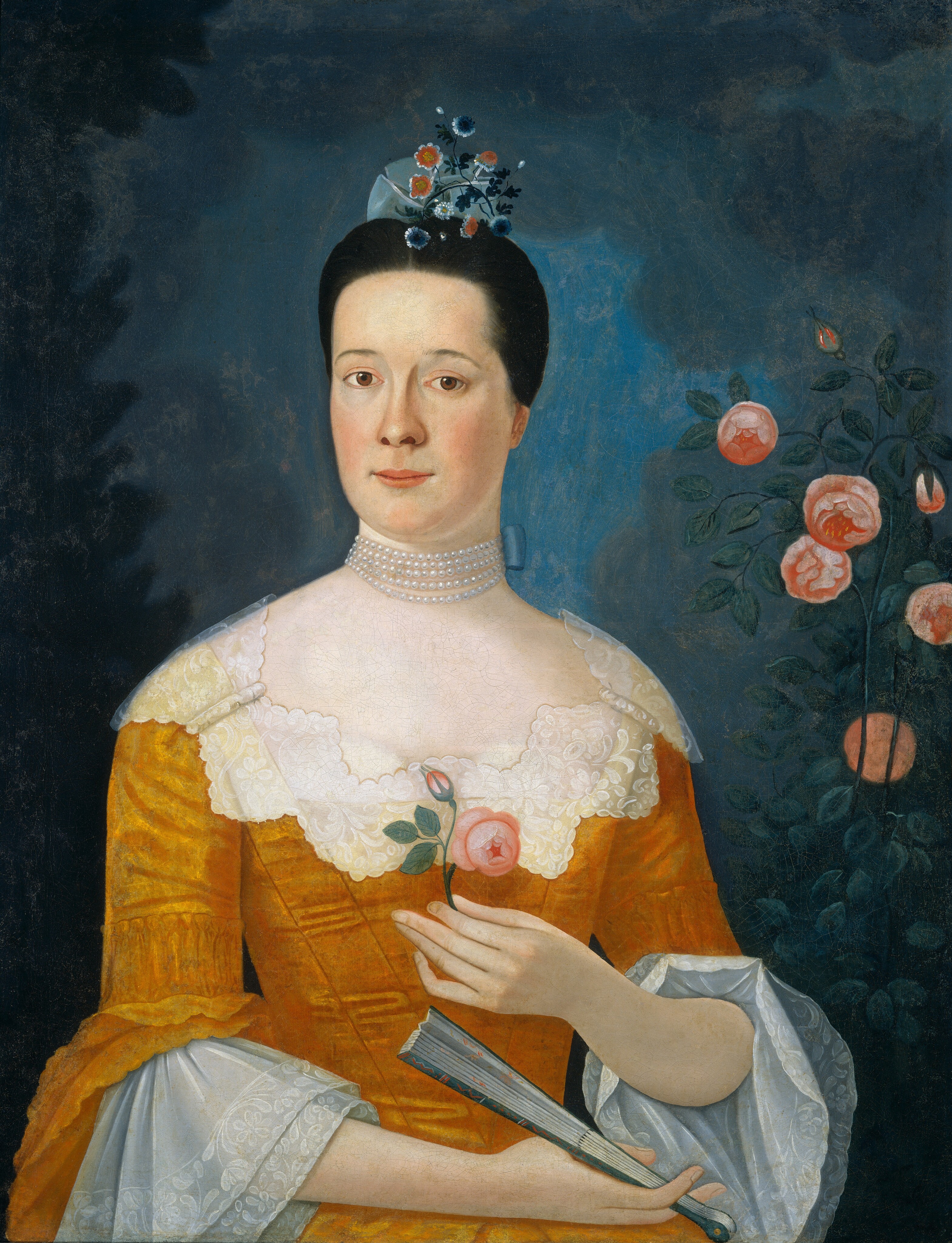 The image shows a woman's upper body from the waist up. She faces slightly to her right but looks directly at the viewer. She has a smooth complexion, bright eyes, and a slight smile. Her dark hair is neatly styled back and adorned with a headpiece of small flowers and ribbons. She wears a mustard-colored gown with lace details, including lace ruffles at the sleeves. She holds a pink rosebud in her right hand and a closed fan in her left hand. A multi-strand pearl necklace is visible around her neck. The background is dark blue with soft pink flowers nearby.