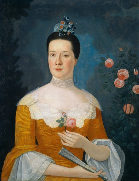 The image shows a woman's upper body from the waist up. She faces slightly to her right but looks directly at the viewer. She has a smooth complexion, bright eyes, and a slight smile. Her dark hair is neatly styled back and adorned with a headpiece of small flowers and ribbons. She wears a mustard-colored gown with lace details, including lace ruffles at the sleeves. She holds a pink rosebud in her right hand and a closed fan in her left hand. A multi-strand pearl necklace is visible around her neck. The background is dark blue with soft pink flowers nearby.