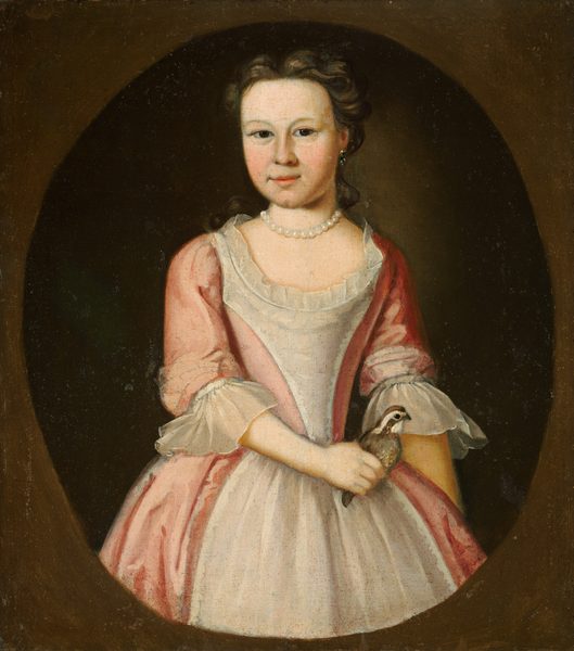 The image shows a young girl from the waist up, slightly turned to her left, with a calm expression. She has fair skin, rosy cheeks, dark eyes, and dark brown curly hair. She is wearing a pink gown with lace-trimmed sleeves, a ruffled neckline, and a string of pearls around her neck. She is holding a small bird in her right hand. The background is dark and oval-shaped, and the portrait is enclosed in a dark circular border.