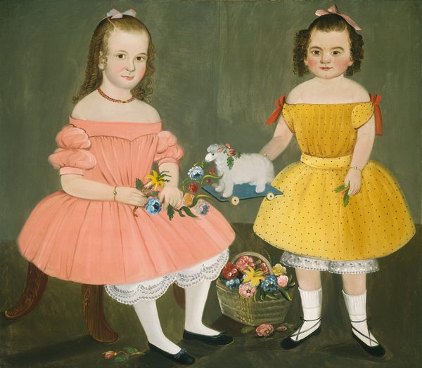 The image shows two young girls standing together. The girl on the left is seated and holding a collection of colorful flowers in her lap. The girl on the right stands holding a toy lamb on wheels. Both girls have curly hair and wear dresses with unique details. The seated girl has a bow in her hair, wears a coral pink dress, and accessorizes with jewelry. The standing girl's dress is yellow with black polka dots and red ribbon accents. Both girls have soft facial features and blush on their cheeks. A woven basket full of flowers is placed between them on the ground. The background is simple and dark.