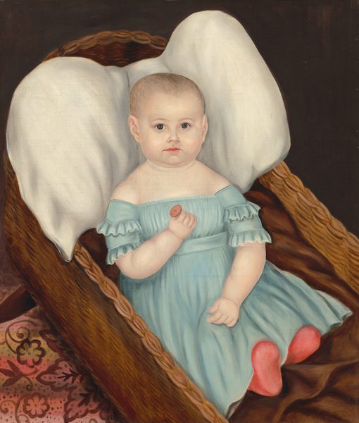 The image shows a baby lying in a wicker cradle. The baby has a round face, light skin, large eyes, sparse light hair, and is wearing a blue dress. The baby is holding a small object in its left hand and is wearing red fabric on its feet. The cradle has a white pillow behind the baby's head and dark brown fabric underneath. The background has a dark area and a patterned floor with floral designs in shades of red and pink.