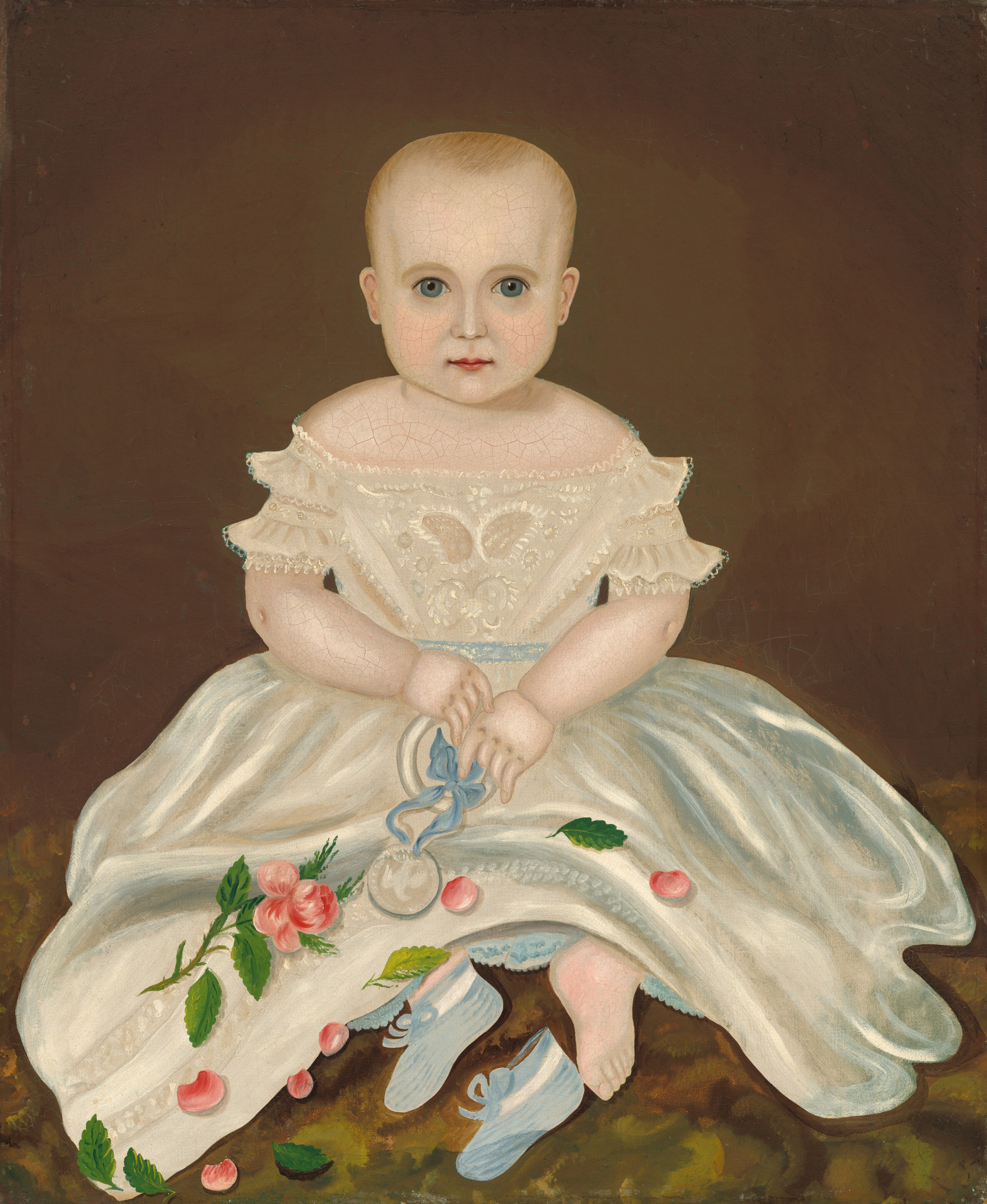 The image shows a seated infant facing forward. The child has a round face with blue eyes and light hair. They wear a white dress with lace and embroidery, painted floral designs in pink and green, and hold a round object tied with a blue ribbon on their lap. Blue shoes are placed next to their bare feet. The background is a plain brown color.