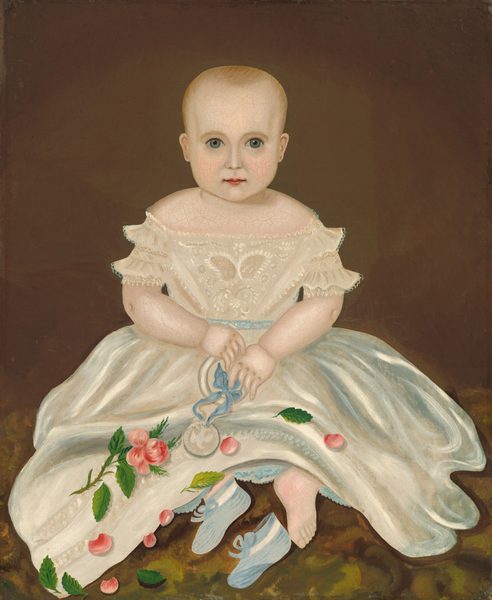 The image shows a seated infant facing forward. The child has a round face with blue eyes and light hair. They wear a white dress with lace and embroidery, painted floral designs in pink and green, and hold a round object tied with a blue ribbon on their lap. Blue shoes are placed next to their bare feet. The background is a plain brown color.