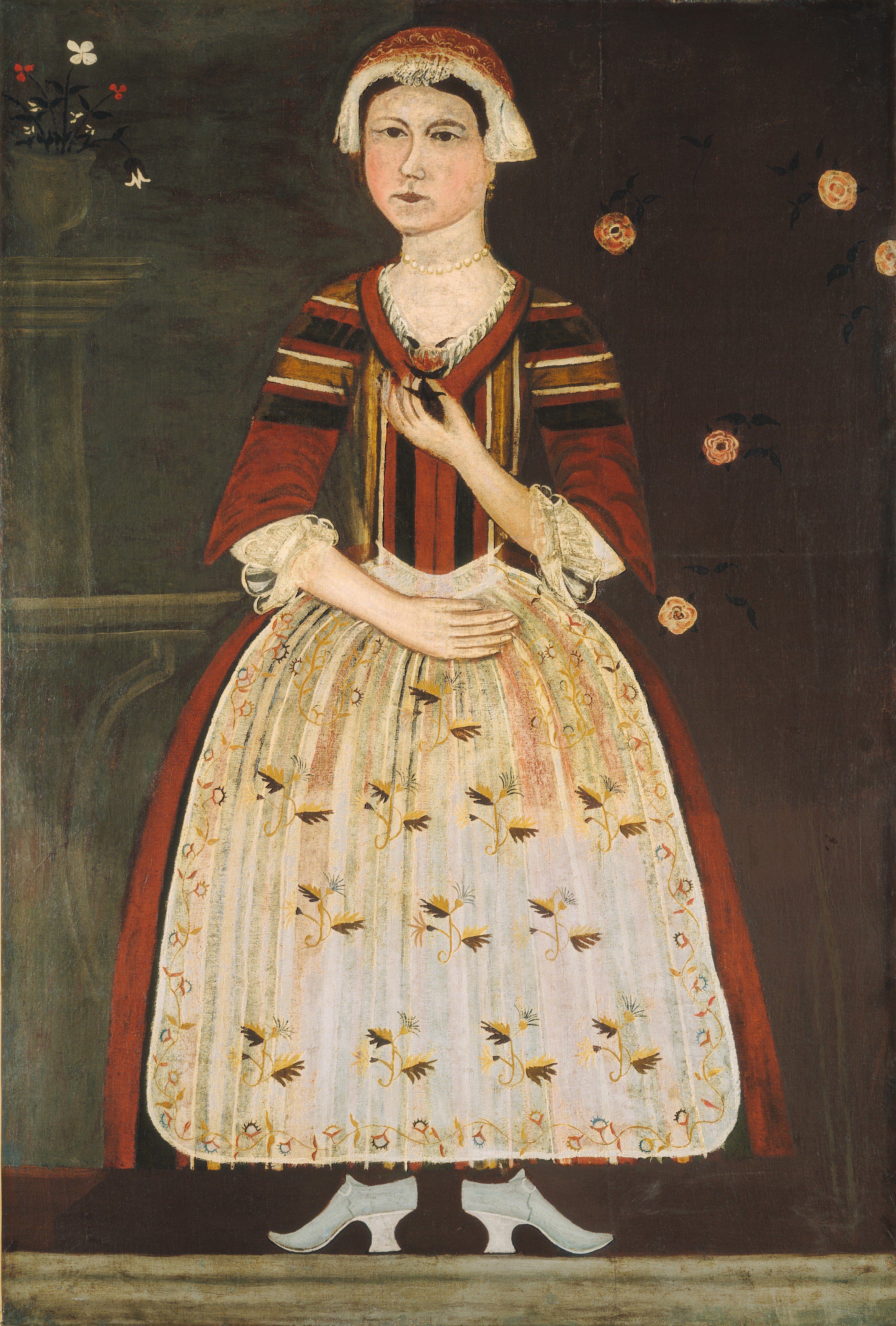 The image shows a woman in a full-length portrait. She has a pale complexion, arched eyebrows, a small mouth, and dark eyes. She wears a cap, a red and gold dress with white lace detailing, and floral patterns. Her shoes are visible beneath the dress, and she wears a pearl necklace. She holds a dark object in one hand and has the other hand on her waist. The background is dark with flowers on the left and a pattern of roses on the right.