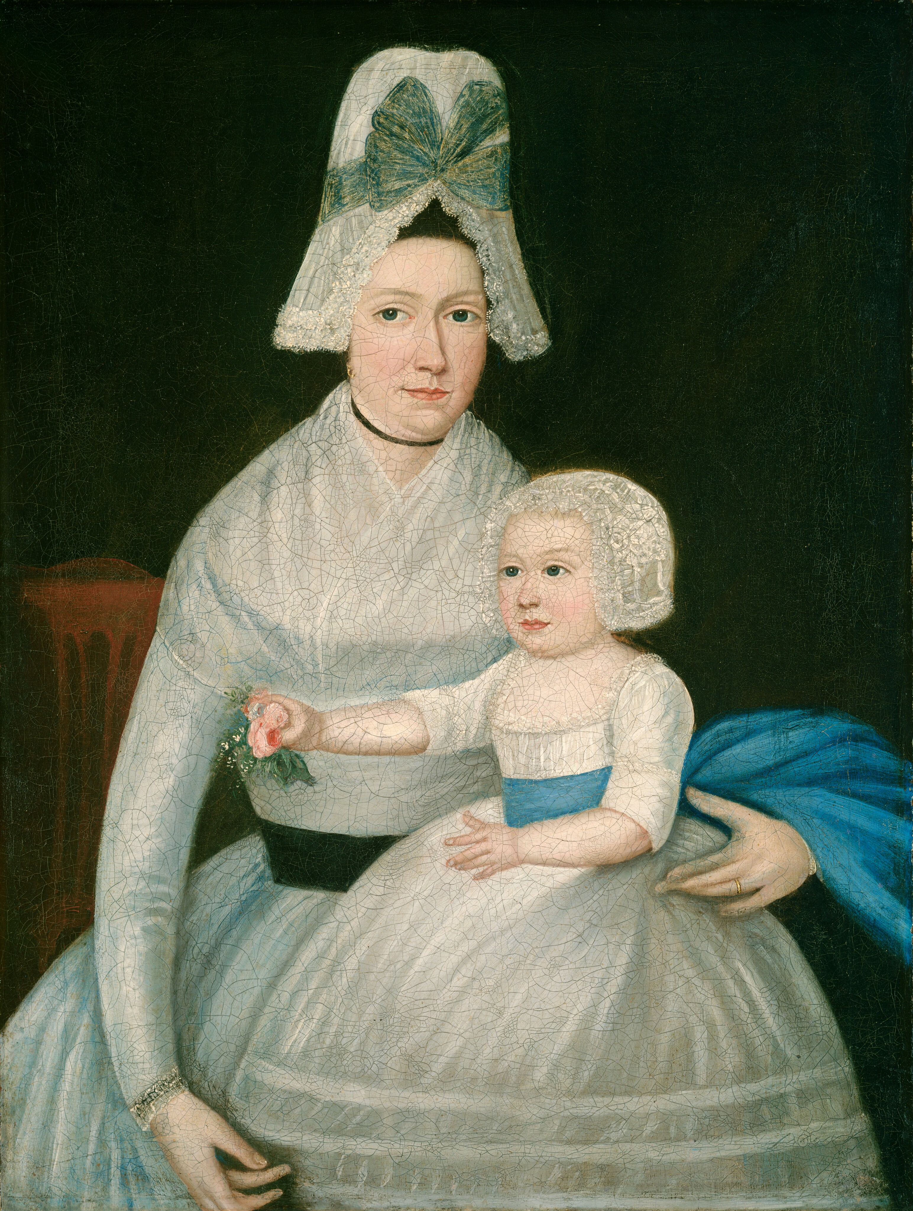 The image shows a portrait of a woman seated with a young child on her lap. The woman is holding the child securely with one arm wrapped around the child and the other holding a small flower. The woman has a fair complexion, an oval face, straight dark eyebrows, light-colored eyes, and is wearing a bonnet with lace and a large blue ribbon. She is dressed in a light-colored gown with a high neckline, a black choker, and a wide black belt. The child, also fair-complexioned, is wearing a dress with a blue sash and a lacy bonnet. They sit against a dark background.