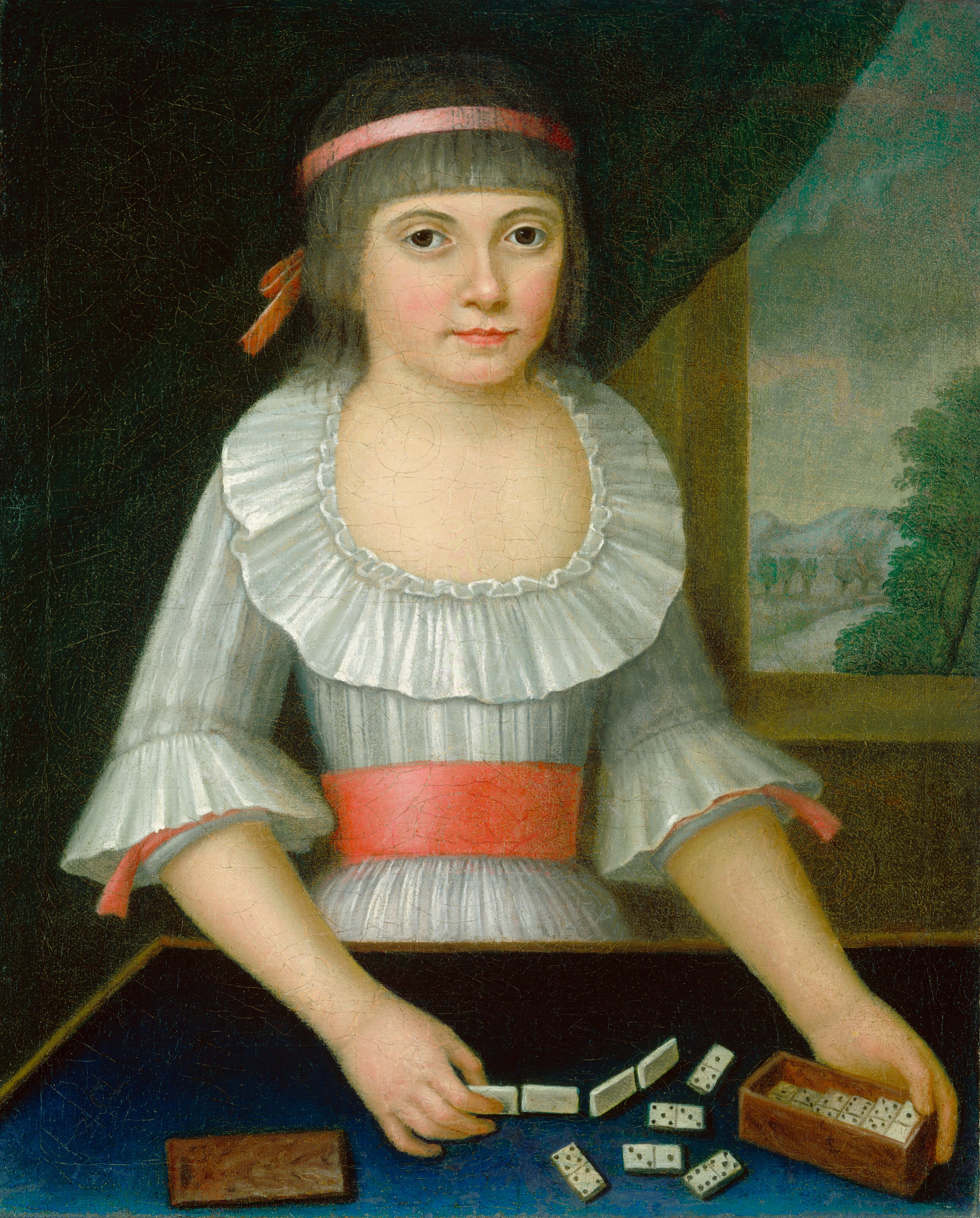 The image shows a young person sitting at a table, holding a box in one hand and arranging dominoes with the other. They have short brown hair with a straight fringe and are wearing a white dress with wide sleeves and a pink sash. The person has fair skin, dark eyebrows, and dark eyes. The background includes a window showing trees and a partly cloudy sky.