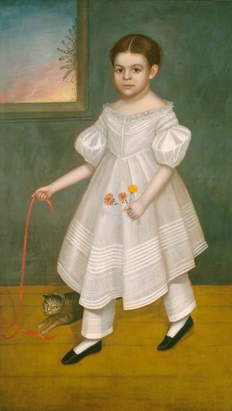 The image shows a full-body portrait of a young child standing indoors. The child faces forward with a neutral expression, looking directly at the viewer. They have neatly styled brown hair parted in the middle. The child wears a white dress with lace detailing at the collar and puffed short sleeves, over layered pantaloons. The dress has intricate patterns along the hem. They hold a bunch of flowers in their left hand and a red ribbon in their right hand, which trails down to the floor. A small cat playfully touches the end of the ribbon near the child's feet. The child wears white socks and black shoes. The background consists of a green-tinted wall and wooden floor, with a window on the left showing a glimpse of the sky with foliage and a warm sunset hue.