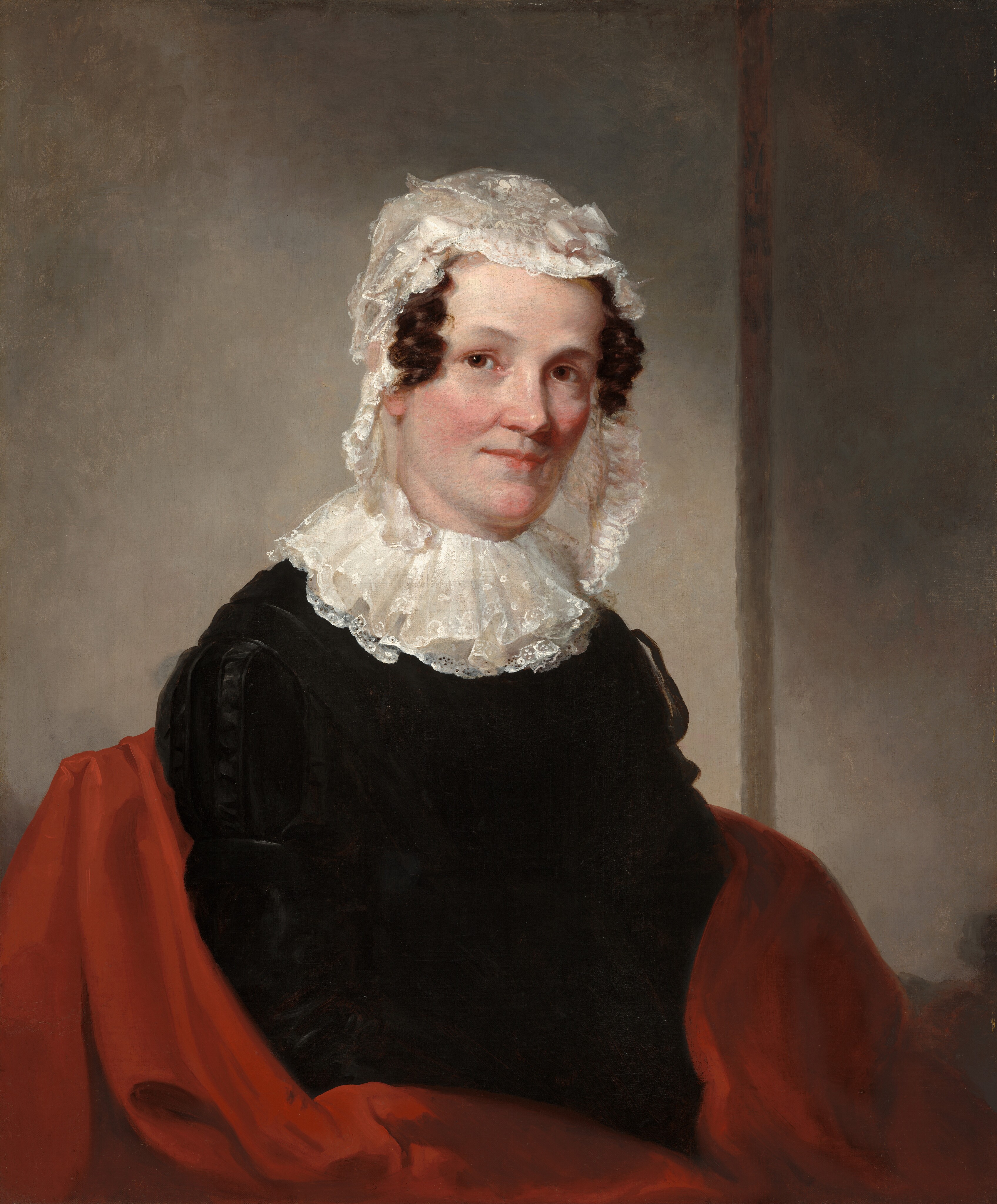 The image shows the upper half of a seated woman, angled slightly to the side but facing forward. She has fair skin, rosy cheeks, dark brown curly hair, and wears a white lace cap with a lace collar over a dark garment. A red fabric is draped around her shoulders. The background is plain with an indistinct vertical line to the side.