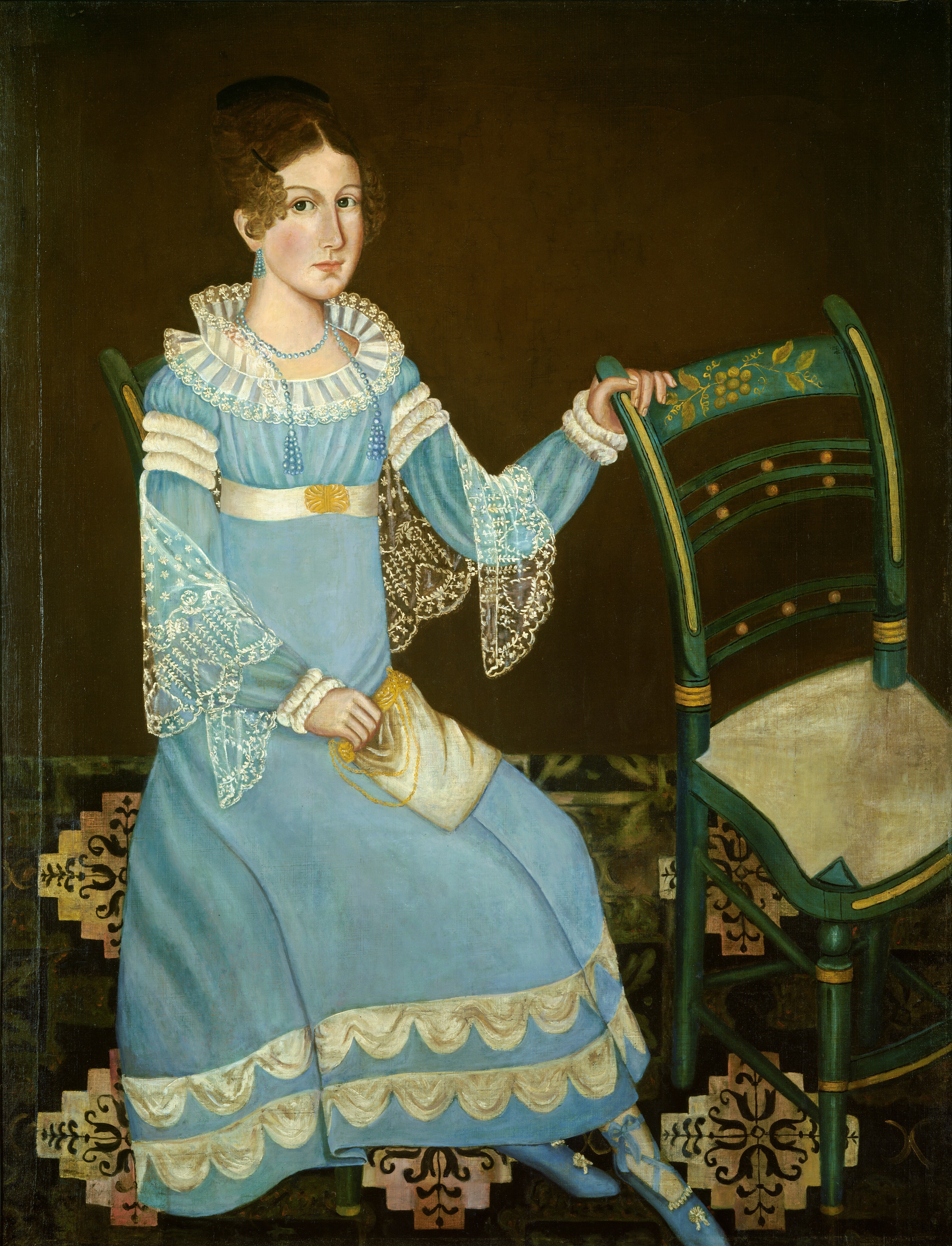 The image shows a person seated on a chair. They face slightly sideways, with one hand on the back of the chair. They have fair skin, dark eyes, and dark curly hair styled up with curls around the forehead. The person is dressed in a light blue dress with ruffled sleeves and a ruffled collar. The dress has white lace details, a decorative belt with a gold buckle, and is paired with blue stockings and shoes with floral detailing. They hold a small white bag or purse and wear earrings and a necklace made of blue beads. The background is plain brown with a patterned floor beneath them.