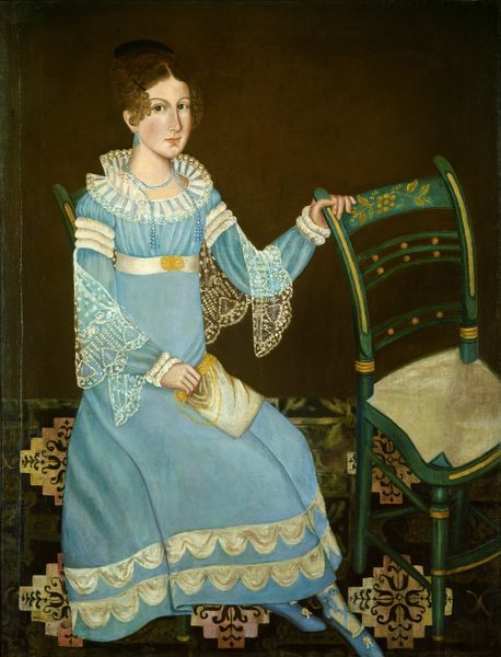 The image shows a person seated on a chair. They face slightly sideways, with one hand on the back of the chair. They have fair skin, dark eyes, and dark curly hair styled up with curls around the forehead. The person is dressed in a light blue dress with ruffled sleeves and a ruffled collar. The dress has white lace details, a decorative belt with a gold buckle, and is paired with blue stockings and shoes with floral detailing. They hold a small white bag or purse and wear earrings and a necklace made of blue beads. The background is plain brown with a patterned floor beneath them.