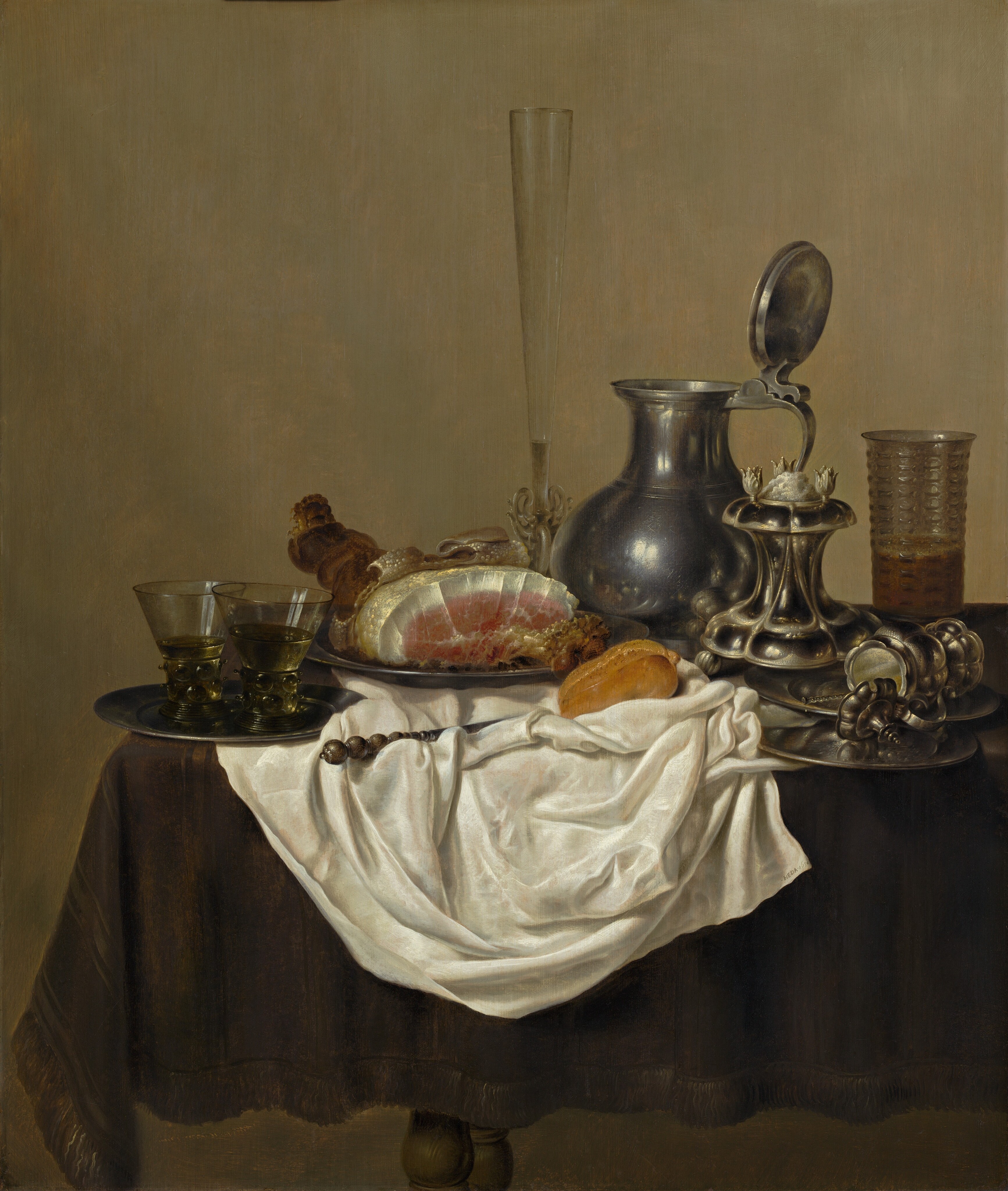 Pewter and silver plates and vessels, a baked ham, roll of bread, and a rumpled, pearl-white cloth are arranged across a table covered with a dark, olive-green cloth in this vertical still life painting. The table extends into the scene from the right and ends just short of the left edge of the composition. The scrunched white cloth falls down the front side of the table, and many of the objects are arranged on it or around its perimeter. To our left, one lip of a pewter plate hangs over the edge of the table and another lies on the white cloth. Two glasses with thick, textured stems and yellow liquid in straight, flaring bowls sit on the plate. Another plate holds a partially carved ham just behind and to our right of the first. The skin of the animal has been peeled back to show the bubbled, white underside. The flesh is white around the edge of the roast and pink at the center. A tall, narrow, clear fluted glass with an intricately woven stem is next to a pewter pitcher with its lid propped open to rest up against the handle. Next, a silver salt cellar has a lobed base that curves in and up to a scallop-edged platform. Another lobed vessel, this one with a handle and a hinged lid, lies open on its side across two overlapping plates at the front of the table. The spiraling end of a utensil protrudes from the vessel. A glass textured with raised dashes is a third filled with amber-colored liquid behind the salt cellar and stacked plates. A roll of bread and a knife with rest in the folds of the white cloth. Turned knobs of the table legs are visible in the gap between the fringe of the green tablecloth and the bottom edge of the composition. The background is an earthy, pale olive green.