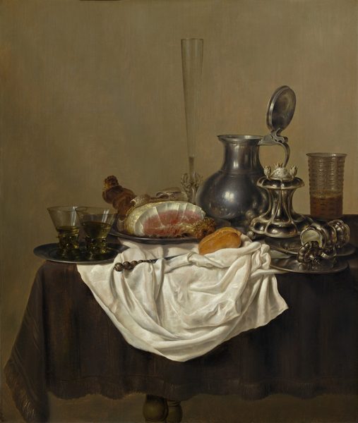 Pewter and silver plates and vessels, a baked ham, roll of bread, and a rumpled, pearl-white cloth are arranged across a table covered with a dark, olive-green cloth in this vertical still life painting. The table extends into the scene from the right and ends just short of the left edge of the composition. The scrunched white cloth falls down the front side of the table, and many of the objects are arranged on it or around its perimeter. To our left, one lip of a pewter plate hangs over the edge of the table and another lies on the white cloth. Two glasses with thick, textured stems and yellow liquid in straight, flaring bowls sit on the plate. Another plate holds a partially carved ham just behind and to our right of the first. The skin of the animal has been peeled back to show the bubbled, white underside. The flesh is white around the edge of the roast and pink at the center. A tall, narrow, clear fluted glass with an intricately woven stem is next to a pewter pitcher with its lid propped open to rest up against the handle. Next, a silver salt cellar has a lobed base that curves in and up to a scallop-edged platform. Another lobed vessel, this one with a handle and a hinged lid, lies open on its side across two overlapping plates at the front of the table. The spiraling end of a utensil protrudes from the vessel. A glass textured with raised dashes is a third filled with amber-colored liquid behind the salt cellar and stacked plates. A roll of bread and a knife with rest in the folds of the white cloth. Turned knobs of the table legs are visible in the gap between the fringe of the green tablecloth and the bottom edge of the composition. The background is an earthy, pale olive green.